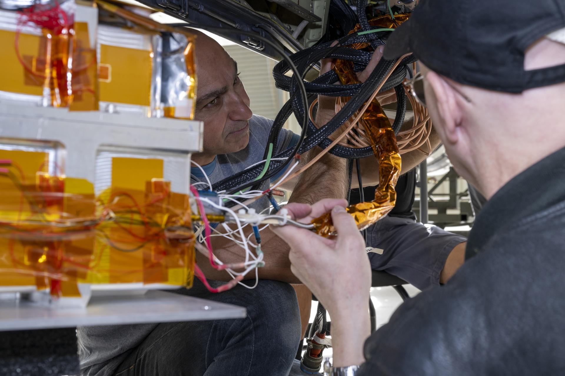 Two men manipulate some entwined cables in the cozy space beneath an aircraft. The cables vary in thickness and color and are attached to a box with bright orange tape enclosing the scientific instrument.