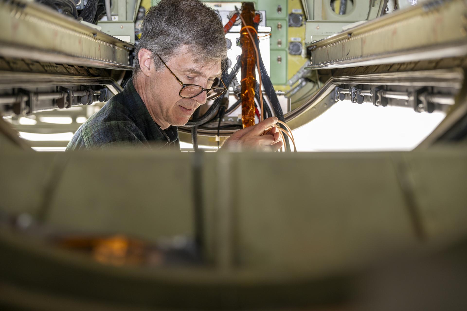 A man’s head and shoulders are just visible as he stands in the open trapdoor to an aircraft’s instrument pod. His hands hold the cables protruding from the interior of the instrument installed on the aircraft, and he focuses on some unseen instrument element beneath the frame of the photo. He has short gray hair and wears glasses that sit at the tip of his nose.