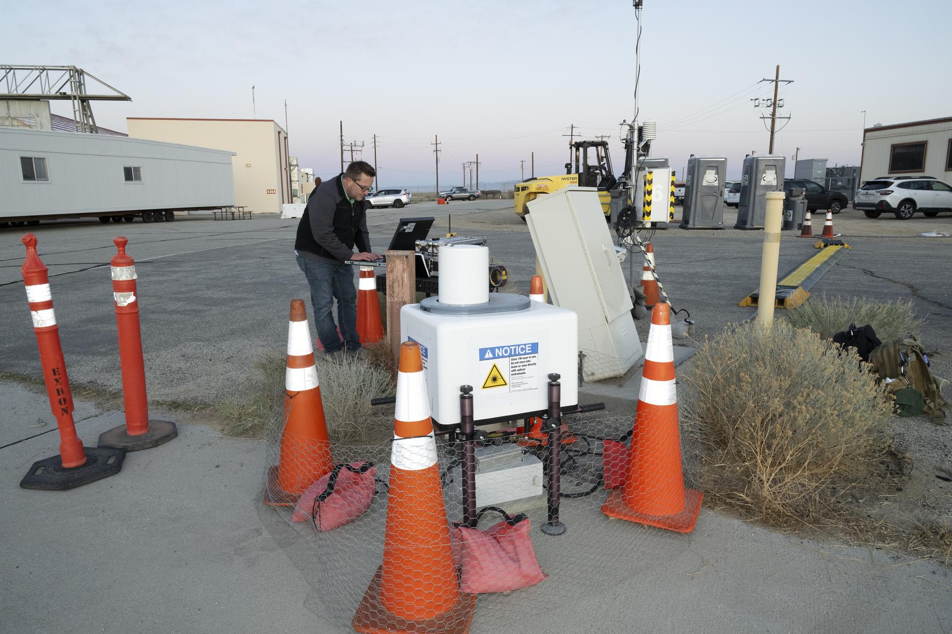 Five orange traffic cones and barriers sit in front of a large white box in the foreground. In the background, a man wearing jeans and a black sweatshirt stands in front of a black laptop. Behind him, there are several cream-colored trailers, other construction equipment, and a few cars.