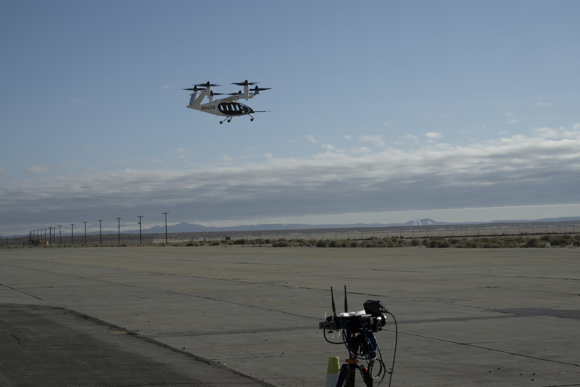 The top of a black tripod with black testing instruments stands in the foreground on a concrete pad with a desert landscape and power lines in the background. A black and white aircraft is in the sky above in the background with blue sky and clouds behind as the aircraft hovers. The aircraft has six black propellors that sit on white arms and connect to the aircraft body, which has black doors and is pod-shaped. The aircraft sits on three small wheels.