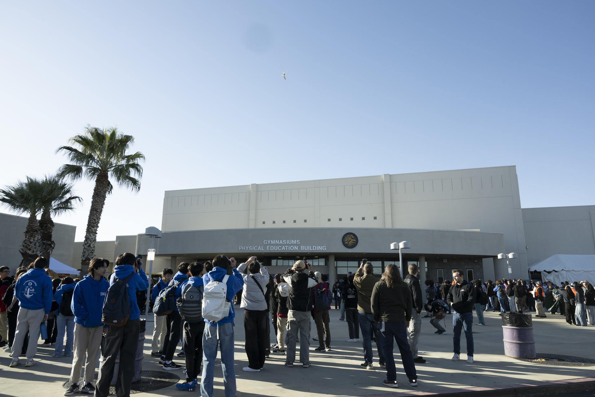 A group of people standing outside a building, watching a jet aircraft fly overhead. The sky is a bright blue and there are palm trees to the side of the building.