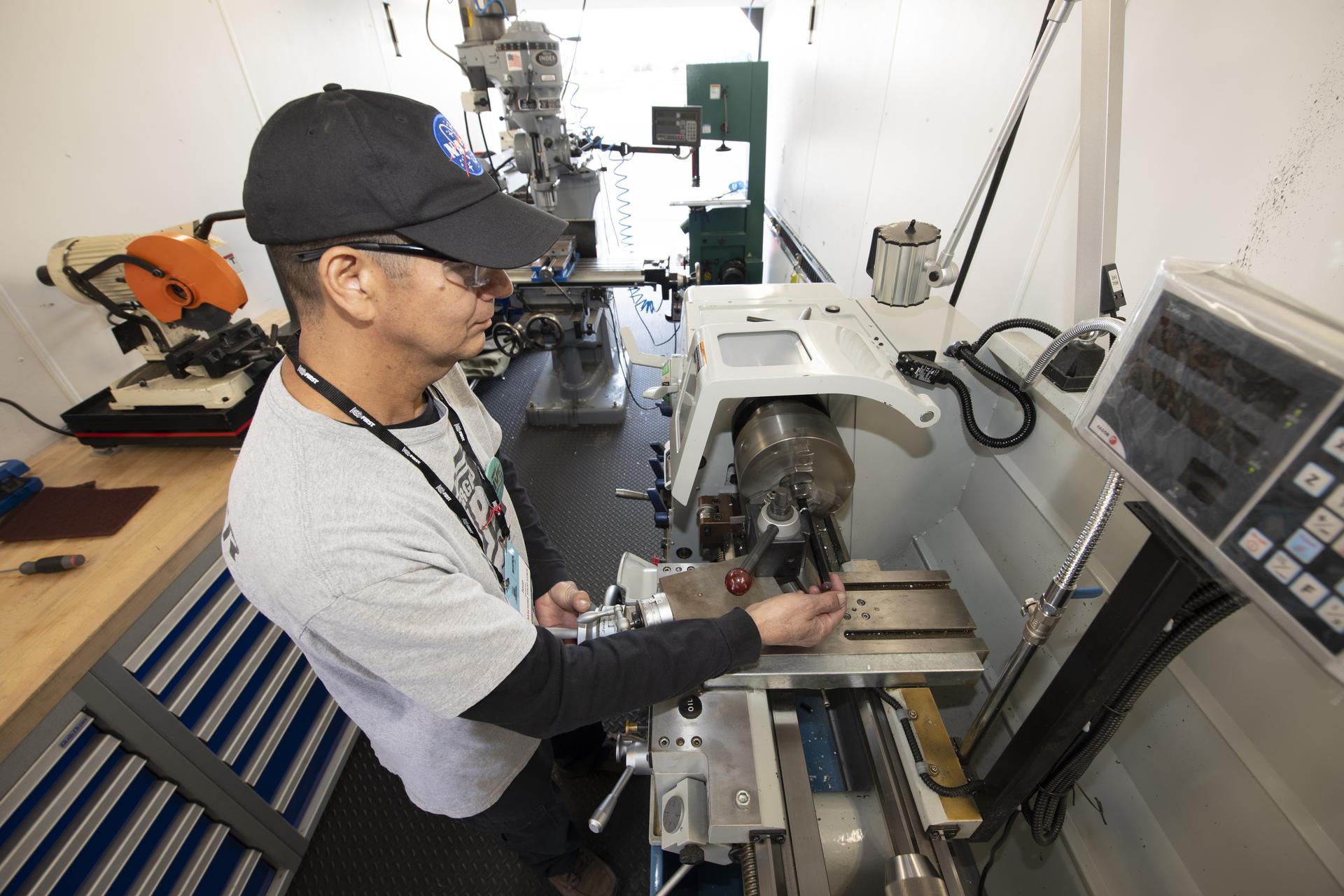 A man wearing a black hat, white shirt, and safety goggles is working on a metal part on a machine. The room is filled with bright light toward the back and the machine is silver.