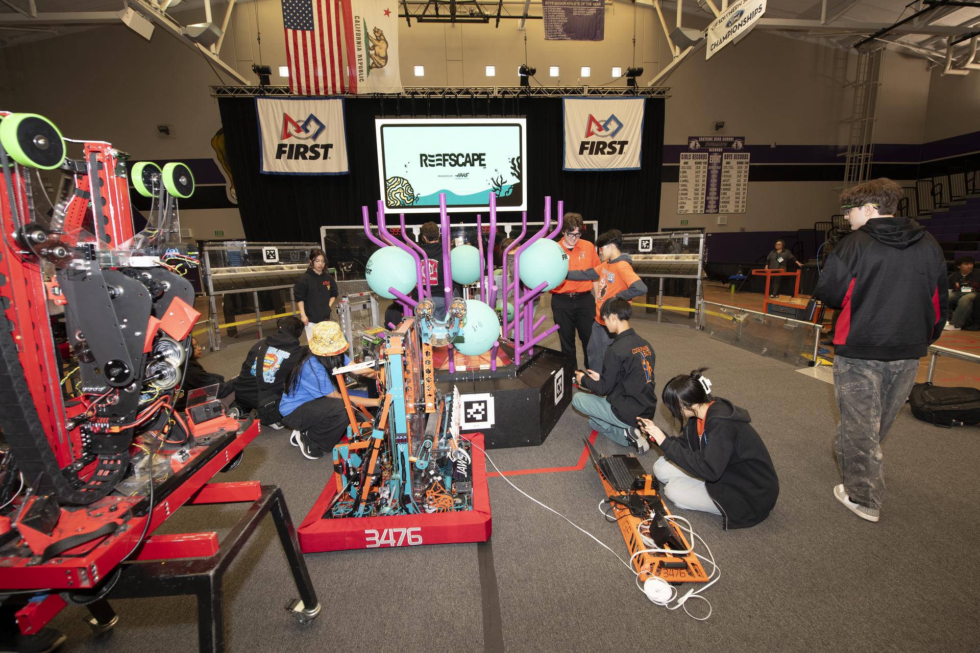A group of students are working on a robot with a red base and lots of gears. Behind the robot is a section on the floor with purple poles holding teal colored plastic.