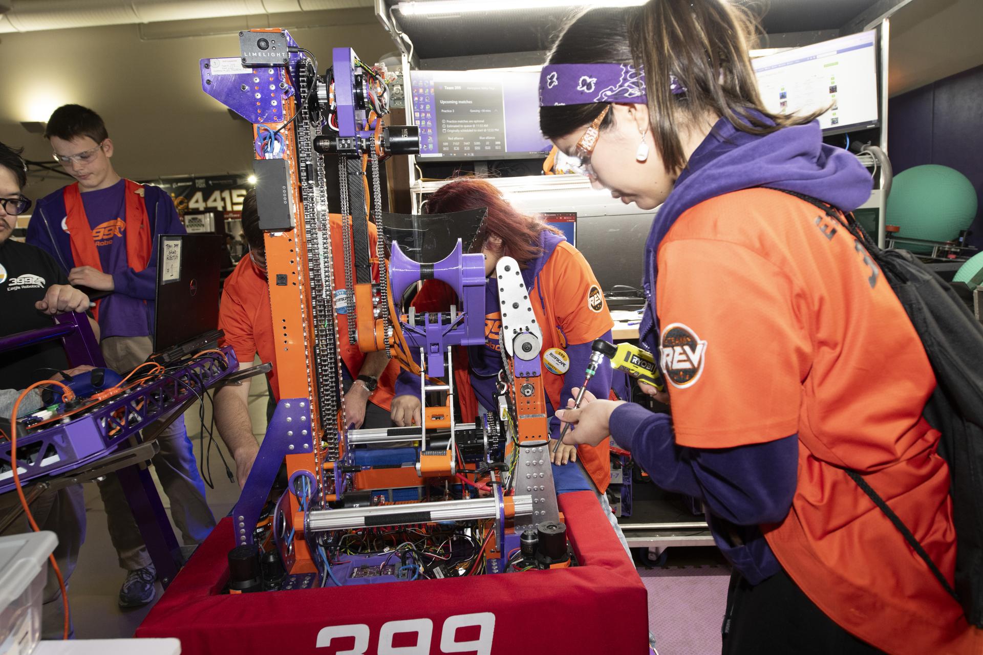 A group of students wearing bright orange jerseys stand around a robot, using tools to adjust and examine its parts