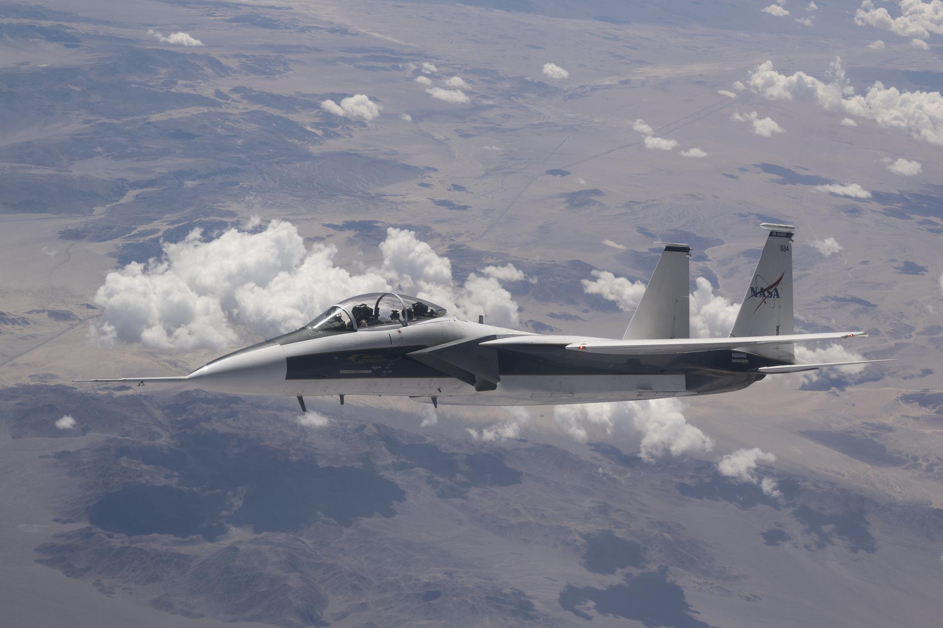 NASA’s F-15D research aircraft flies above scattered clouds with landing gear retracted during a calibration flight near Edwards, California. The aircraft carries a long shock-sensing probe mounted on its nose, and the NASA logo is visible on the tail below the number 884.