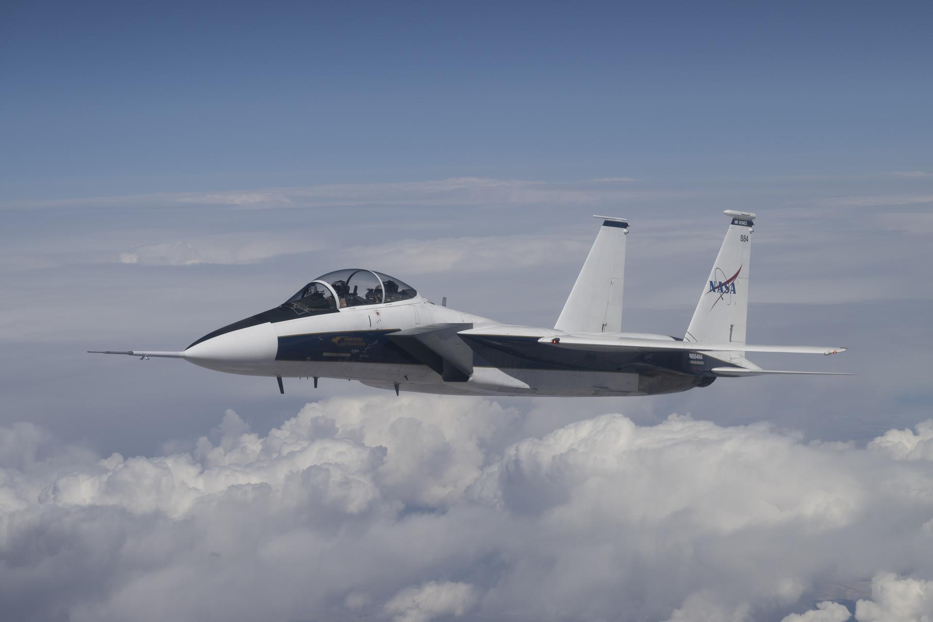 NASA’s F-15D research aircraft flies above scattered clouds with landing gear retracted during a calibration flight near Edwards, California. The aircraft carries a long shock-sensing probe mounted on its nose, and the NASA logo is visible on the tail below the number 884.