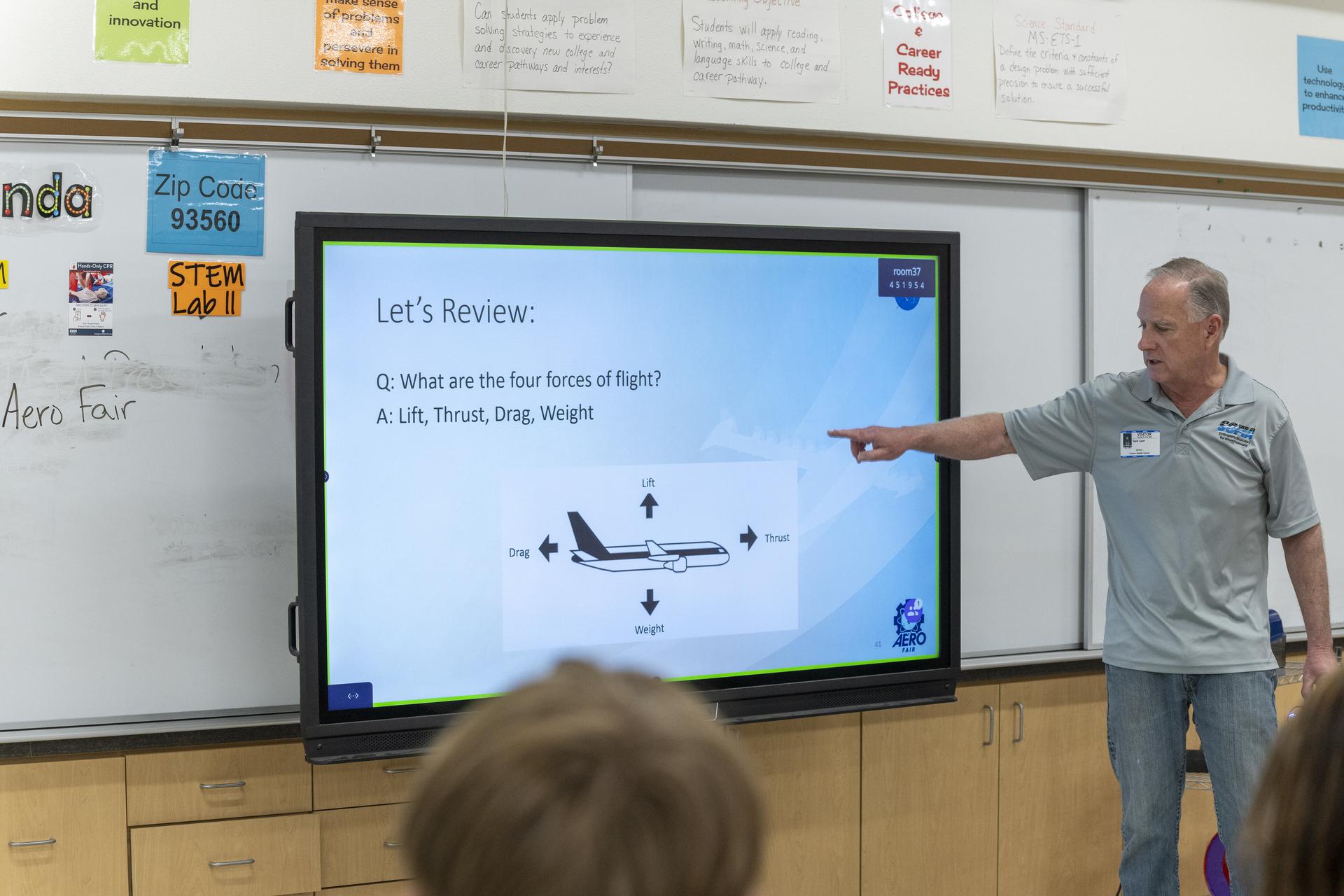 A man wearing a gray shirt is pointing to a screen on a wall displaying an aircraft. Students in the classroom watch the screen and the man.
