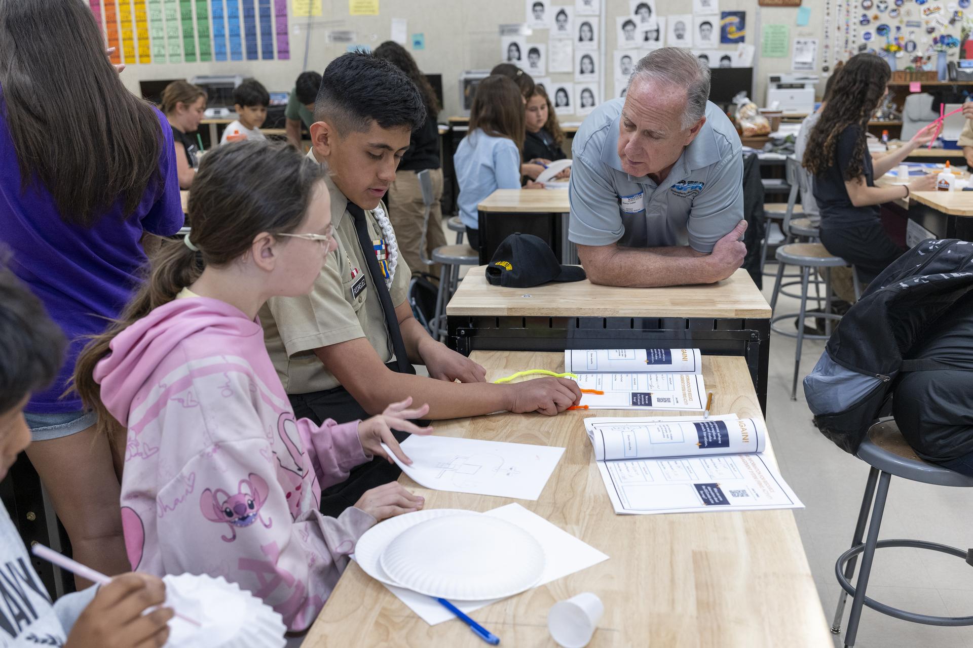 A group of students sitting at a desk in a classroom work on constructing paper airplanes. A man leans on the desk, speaking with the students. Behind the man are other students seated at desks.