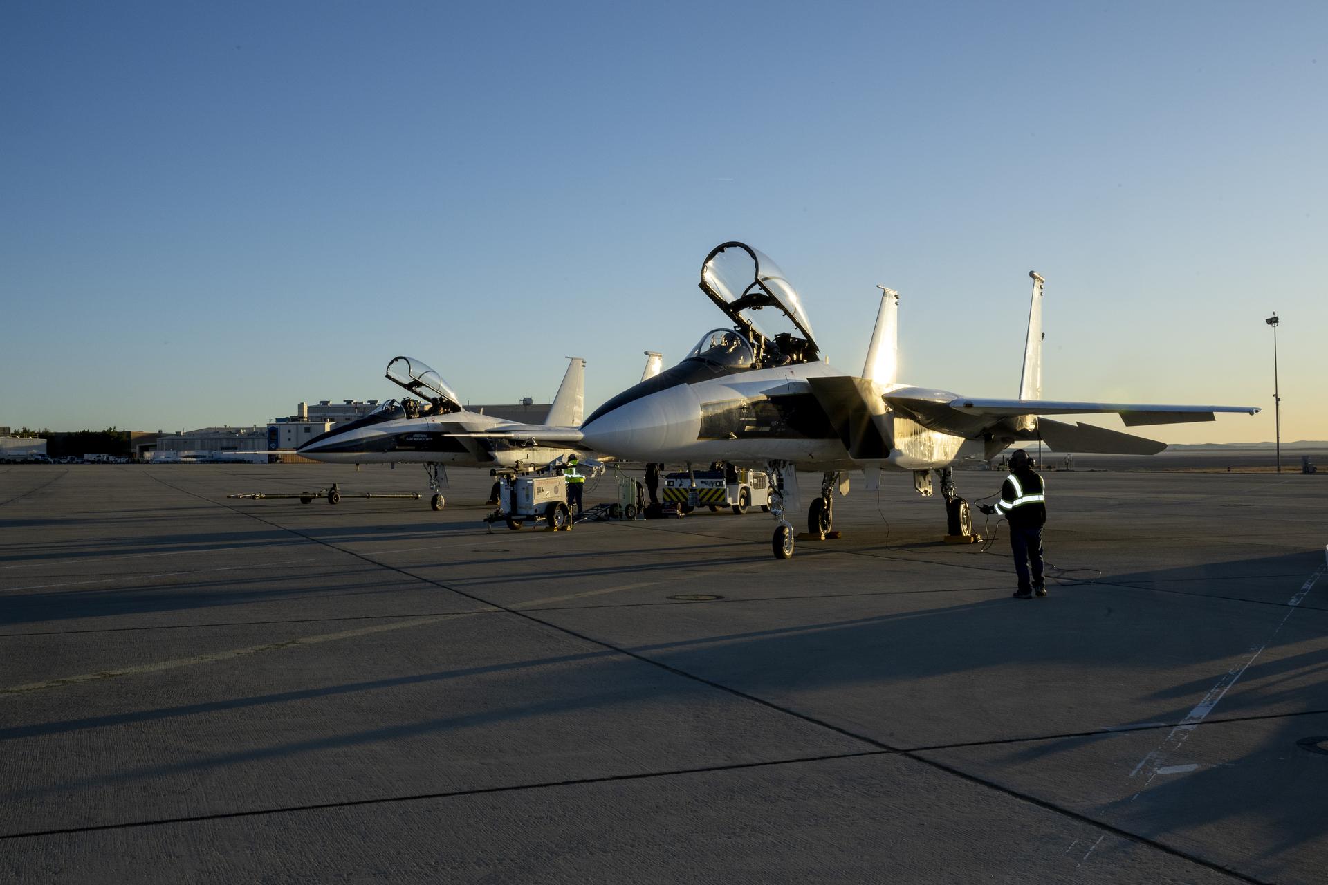 Two NASA F-15 aircraft are parked on the ramp at sunrise at NASA’s Armstrong Flight Research Center in Edwards, California. The aircraft canopies are open with the nose-mounted shock-sensing probes visible. The aircraft face toward the viewer with the sun rising behind them. A single ground crew member wearing a reflective vest stands near the left wing of the closer aircraft. Between the two aircraft is another ground member in a reflective vest working the aircraft tug and fuel cart. The sky above is a bright, light blue.
