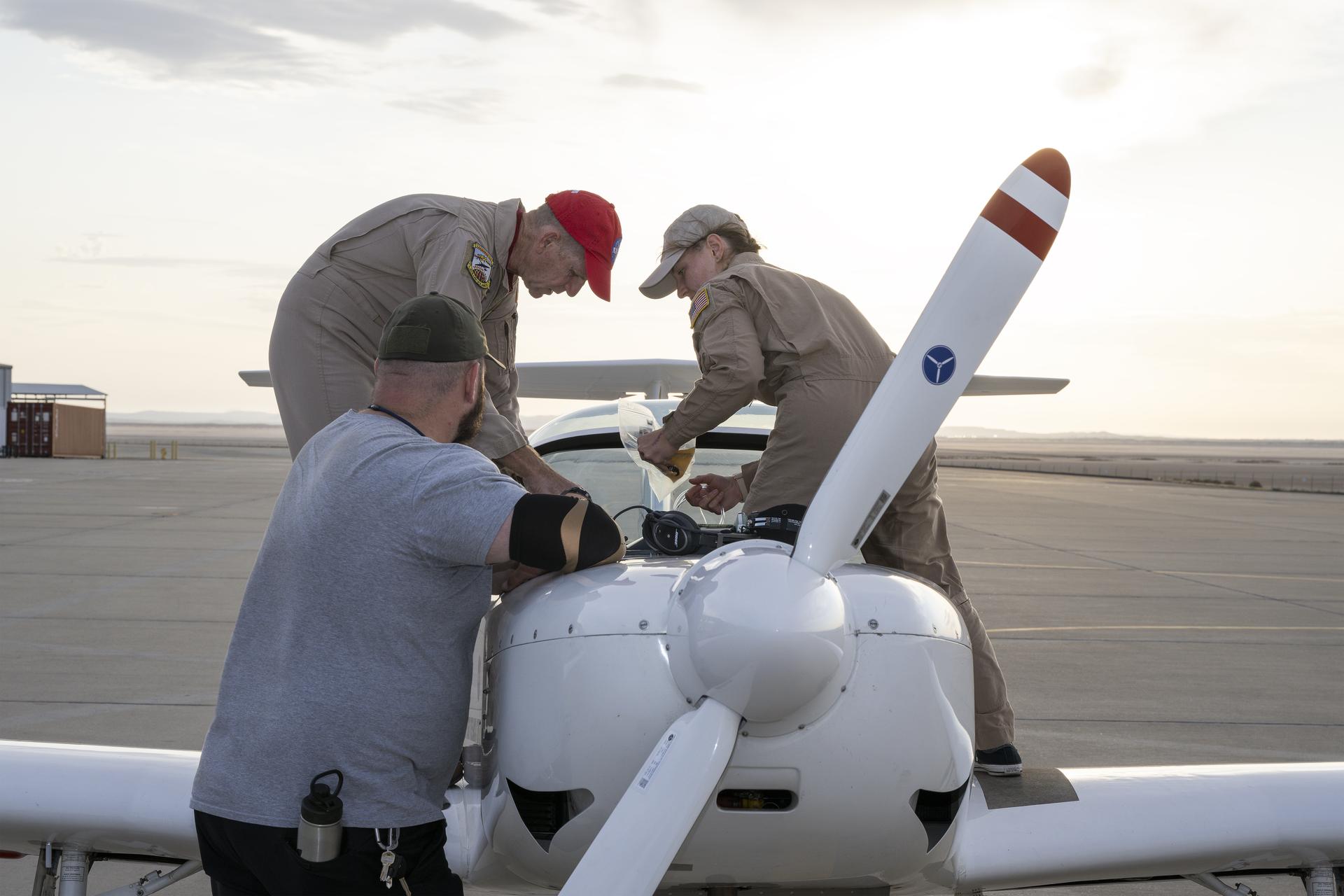 A NASA TG-14 glider aircraft sits on the ramp at NASA’s Armstrong Flight Research Center in Edwards, California. Two pilots stand over the cockpit with the canopy open while the crew chief watches them work. The white glider has red accents on the nose, and the NASA logo is visible on the propeller. The aircraft is parked on a wide, open ramp with desert hills in the distance under a pale sky.