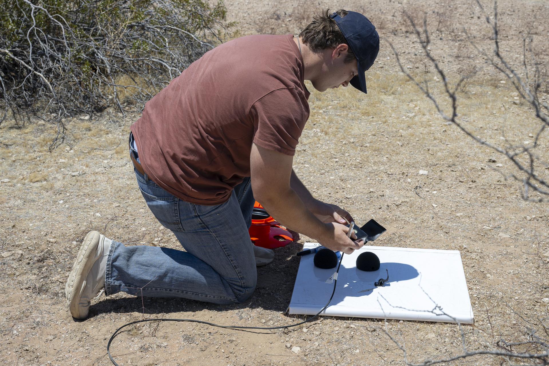 A NASA intern kneels on the desert floor, positioning a microphone on a white platform as part of a ground recording system (GRS) setup. He wears a maroon shirt, jeans, and a dark ballcap. Desert shrubs and dry terrain stretch into the background under bright sunlight. The GRS equipment includes a microphone, foam windshields, and cables for data collection.