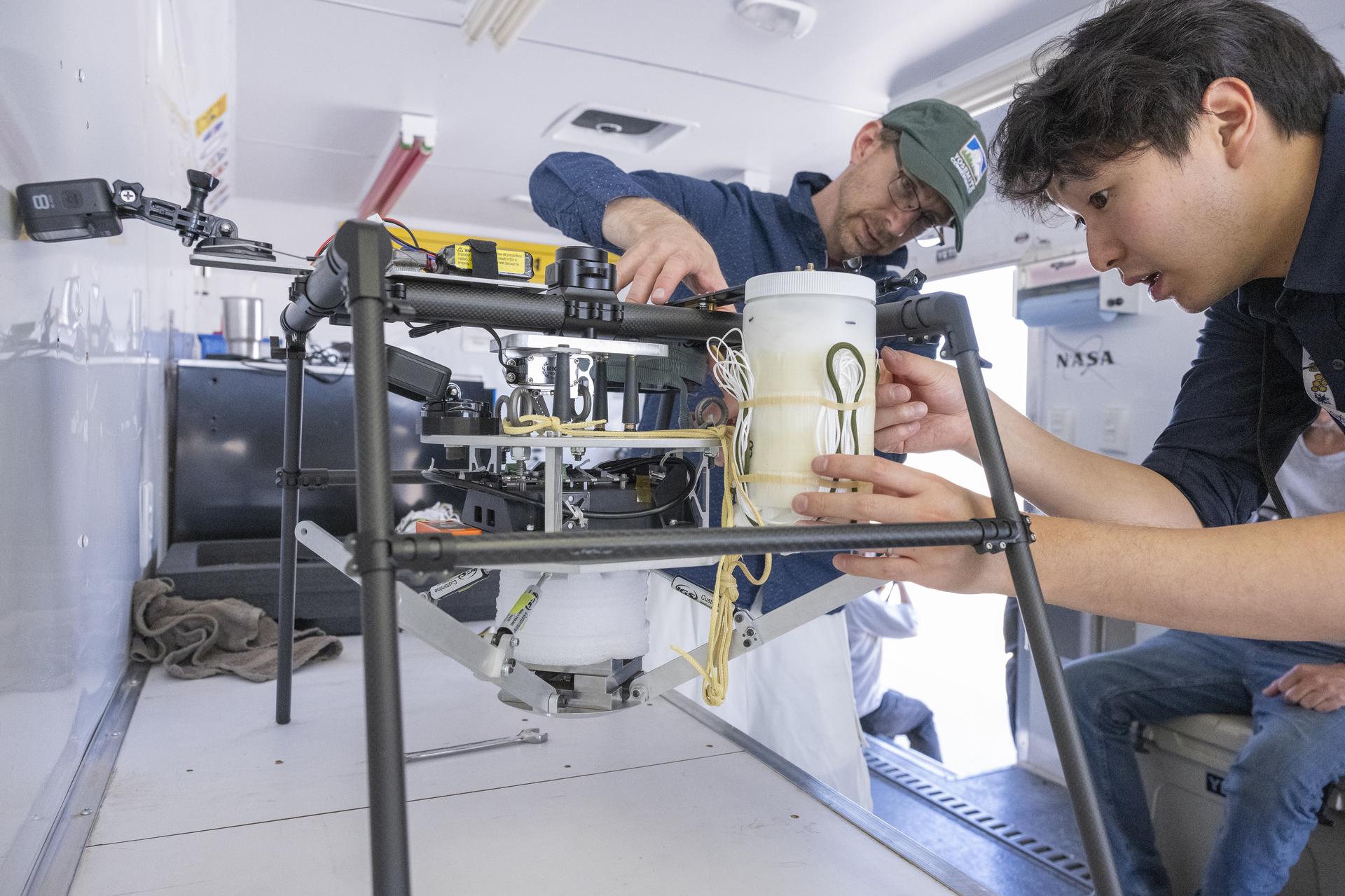 One man secures a cannister containing a parachute to a system that will be carried by an Alta X drone, while another man assists.