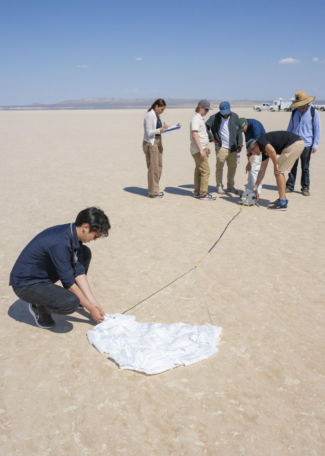 A group of researchers on a barren dry lake examine a capsule after it was released from a drone and carried by an instrumented parachute.