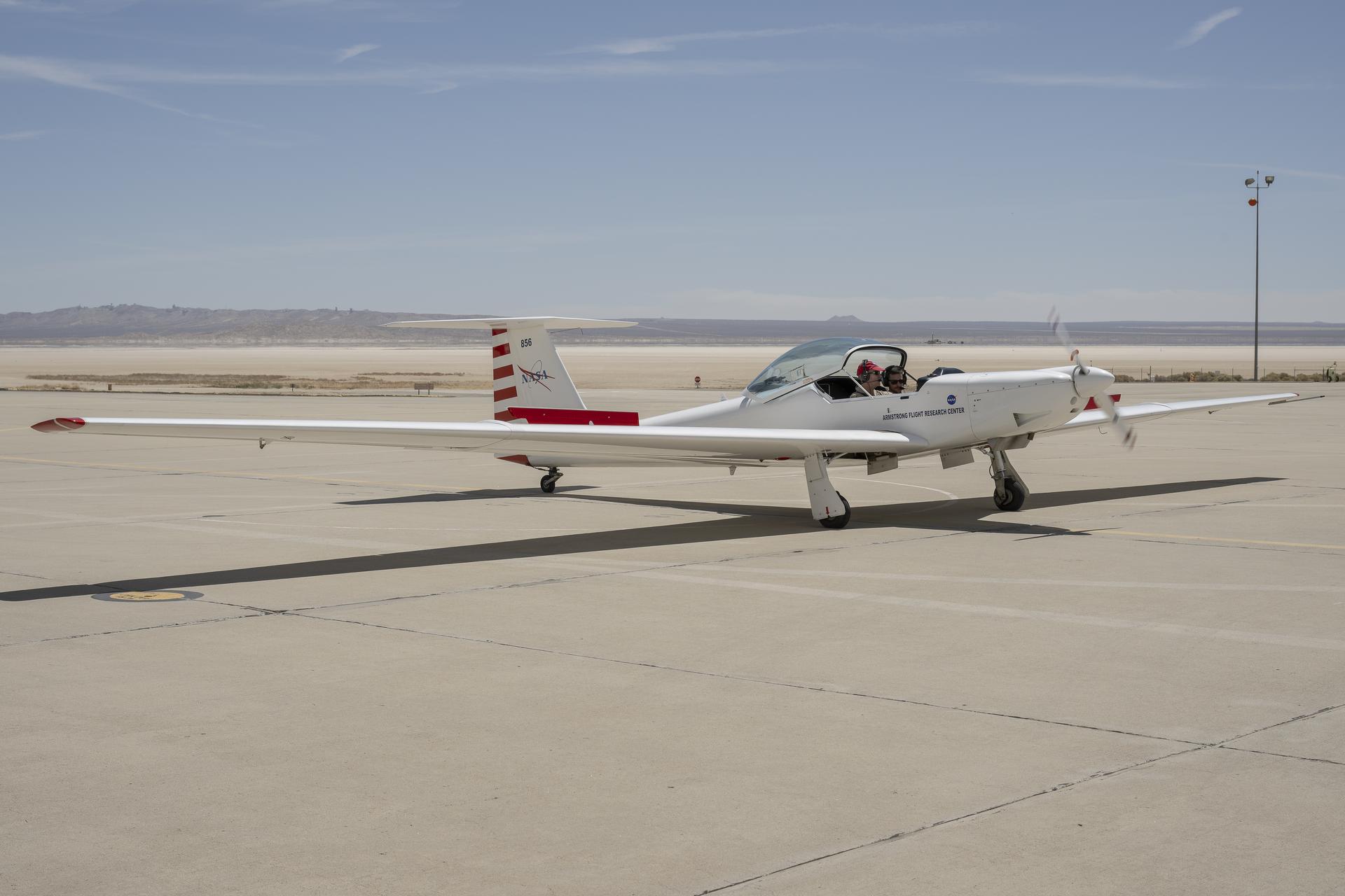 A white airplane with long, thin wings sits on a concrete ramp. The airplane has red stripes on the tail. Two people wearing headphones sit side-by-side inside the cockpit. The glass canopy is open. The aircraft’s front propellor is moving and is blurred in this image by the motion. The background is flat and sand colored with mountains behind.