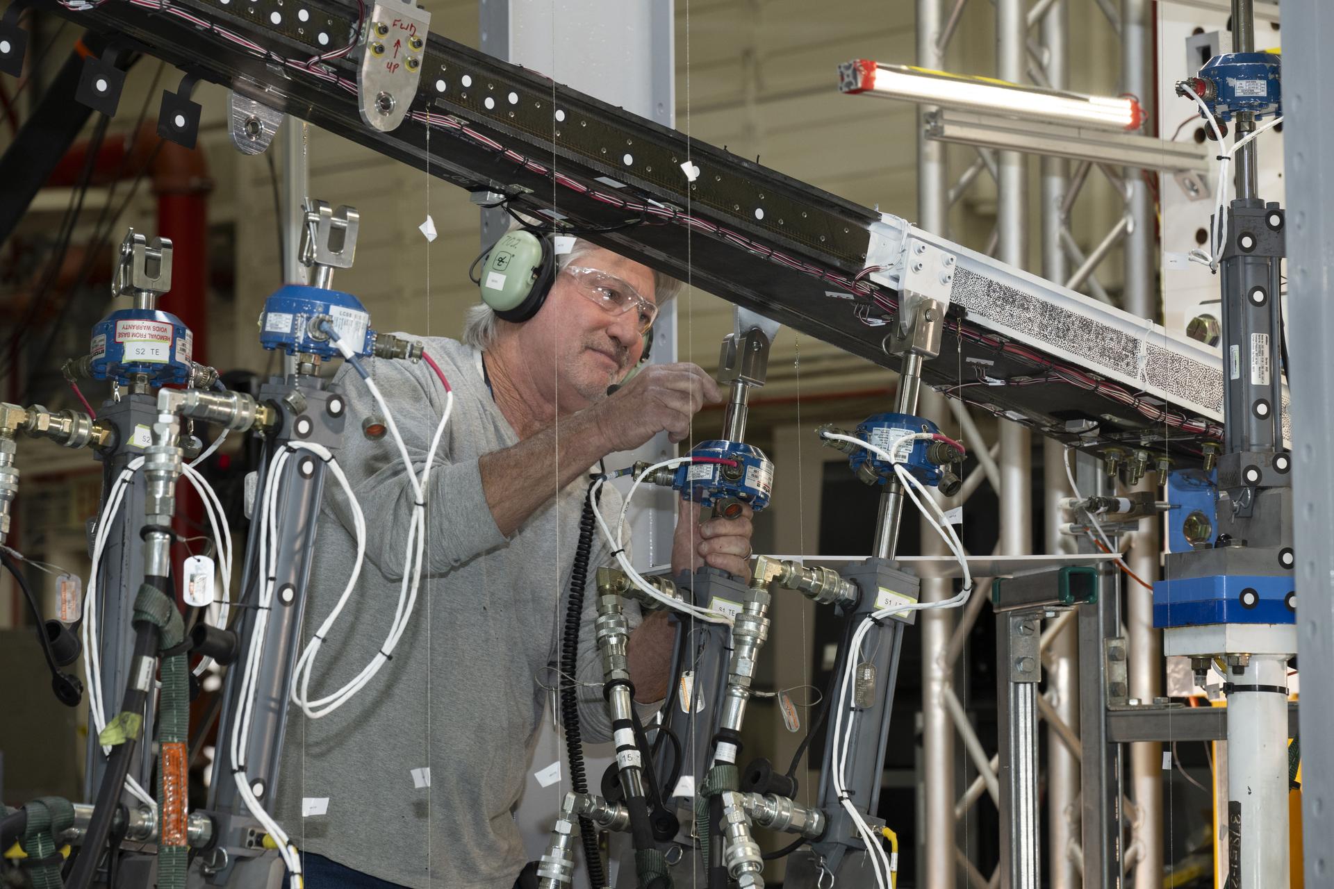 A man wearing ear protection works closely with multiple hydraulic and instrumentation units connected to a large beam mounted on a test structure. Numerous cables, hoses, and measurement devices extend from the setup.