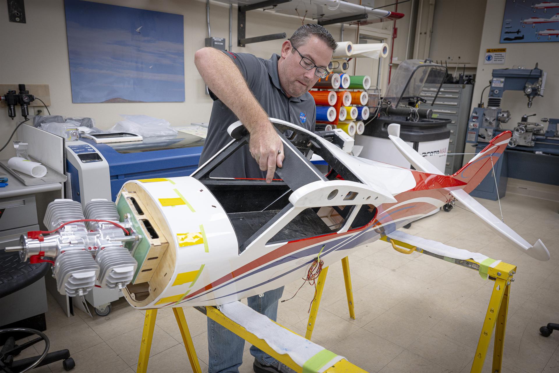A man rotates a subscale aircraft onto its side in preparation for aligning the engine cowl. He will apply yellow tape to guide the cowl’s proper placement during installation.