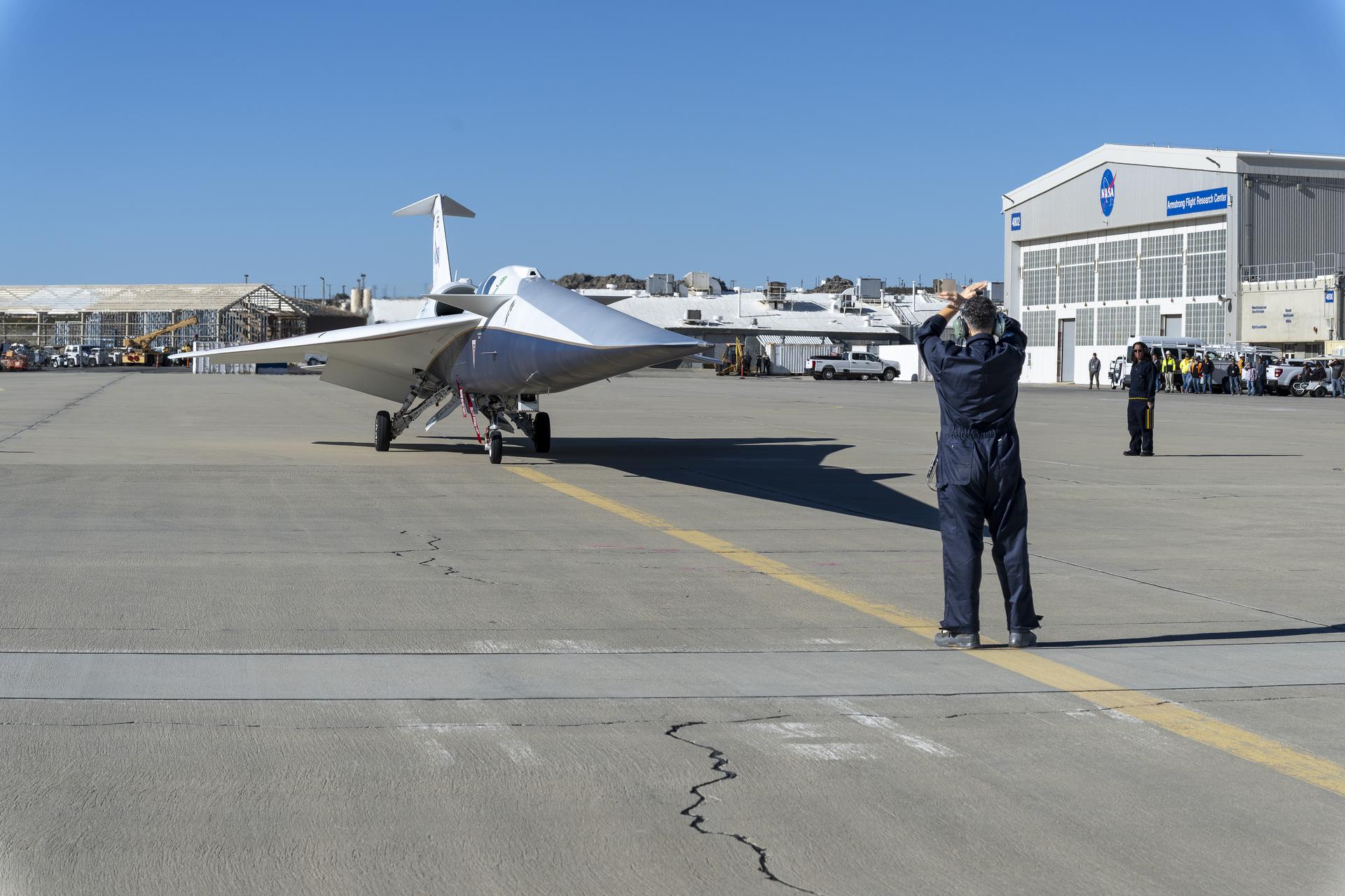 The X-59 pulls onto the ramp at NASA’s Armstrong Flight Research Center. In front of the aircraft, the crew chief stands with arms raised to guide it in, while another flight crew member stands beside the aircraft. An audience is visible in the background near a large hangar.