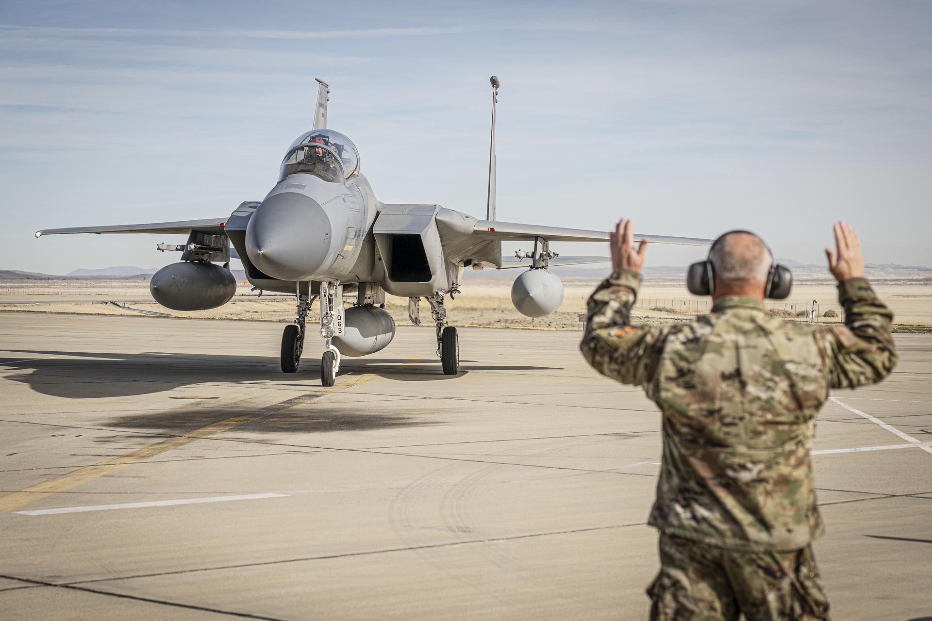 Alt Text: A ground crew member wearing hearing protection raises both arms to guide an F-15 aircraft taxiing on the ramp at NASA Armstrong Flight Research Center.
