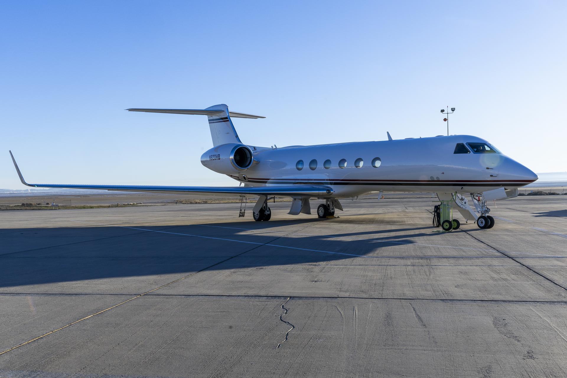 A white airplane sits on a concrete ramp. In view is one of the two engines and one of the two wings. The aircraft has four sets of wheels underneath, six windows, and a three-window cockpit. One red and one blue stripe stretches across the belly of the aircraft.