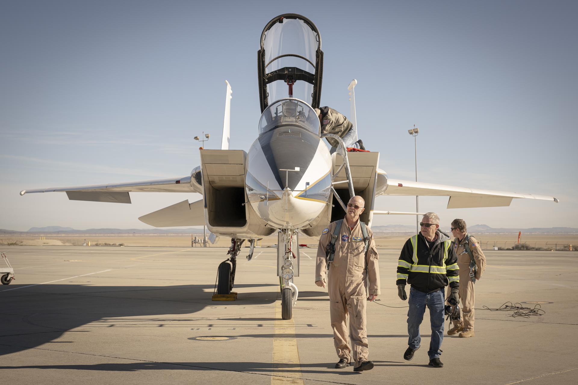 Three men, two wearing tan flight suits, and one wearing a black jacket with stripes of reflective yellow coating, walk to the right side of a jet aircraft. The aircraft faces forward and the canopy is open. Two wheels, intakes, wings, and tails are in view of the white aircraft. The aircraft sits on a concrete ramp with desert sand and mountains in the background.