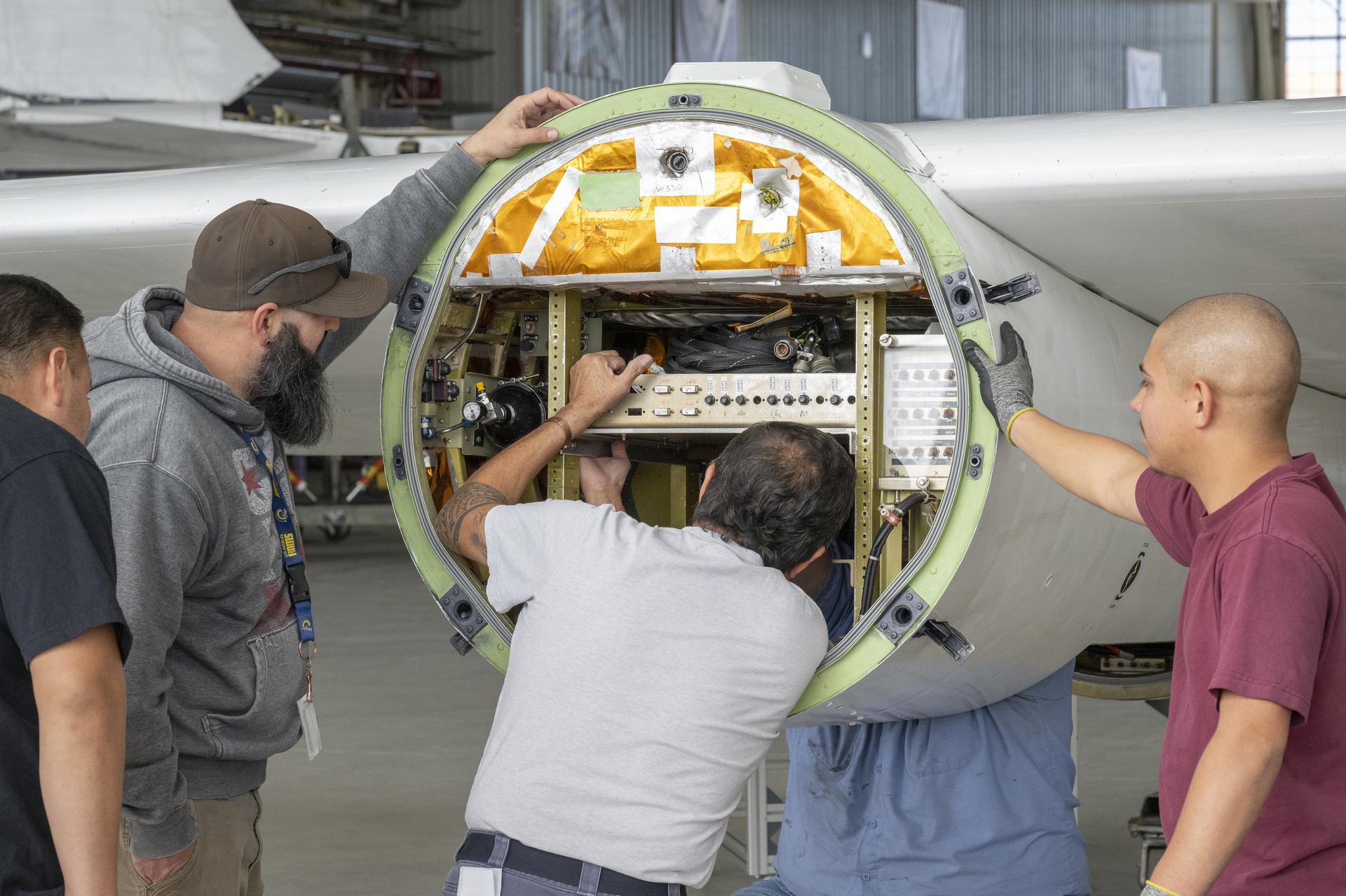 Four men surround a round object attached to an aircraft wing with shelves and wiring inside. One man’s arms are inside of the object, and he is adjusting a gold-colored piece of metal equipment. The other men watch as he works on the hardware.