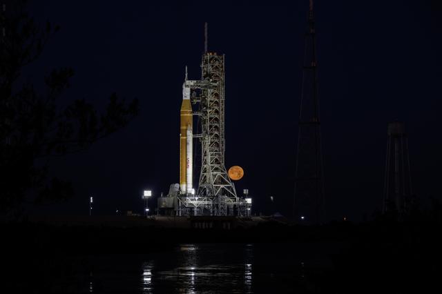 The Moon sets behind NASA’s Artemis II SLS (Space Launch System) rocket and O...
