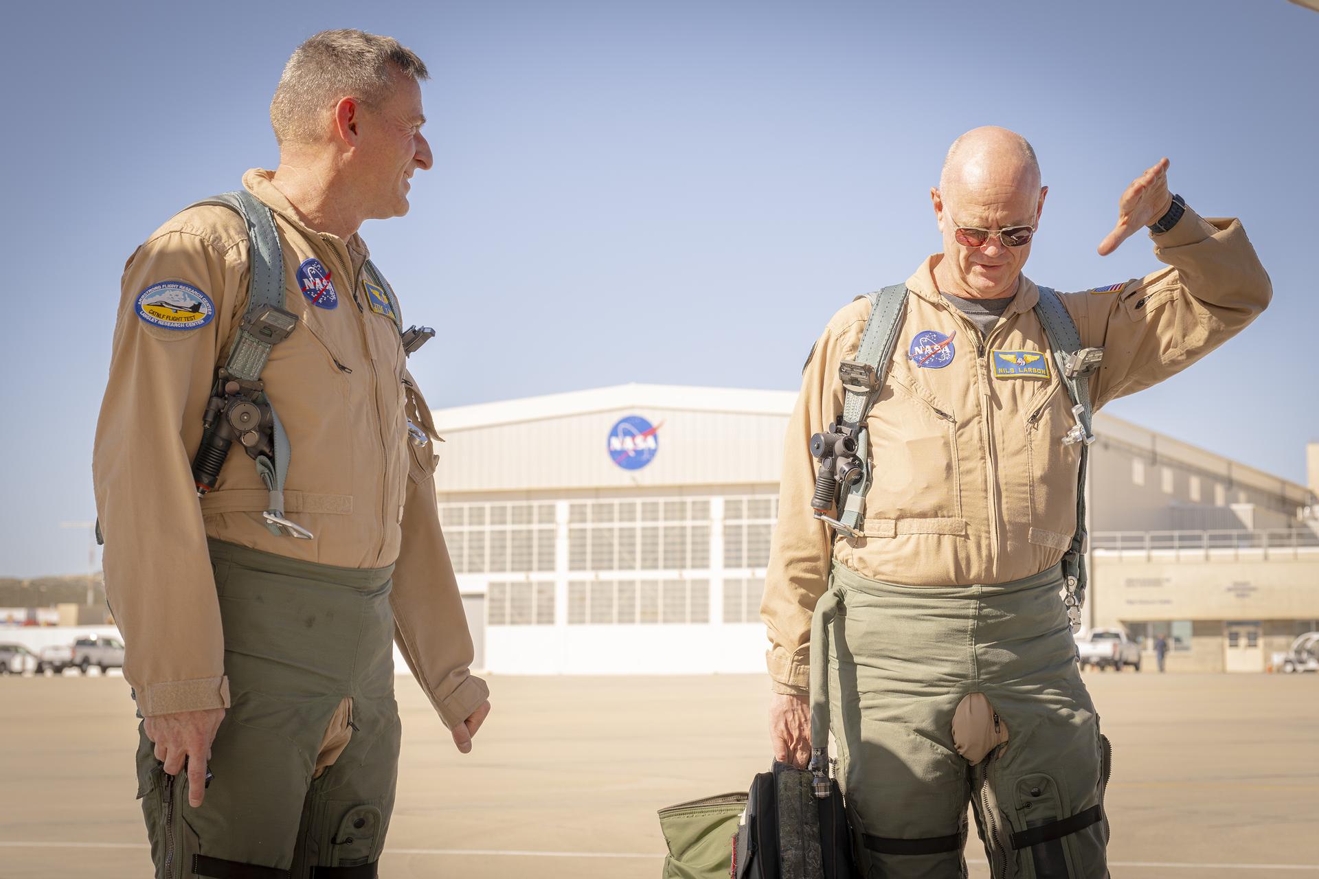 Two men, wearing tan flight suits and green flight gear, walk on a concrete surface away from a building in the background. The man on the left looks at the man on the right as he is moving his left hand in the air. This same man is carrying a bag with flight gear in his right hand.