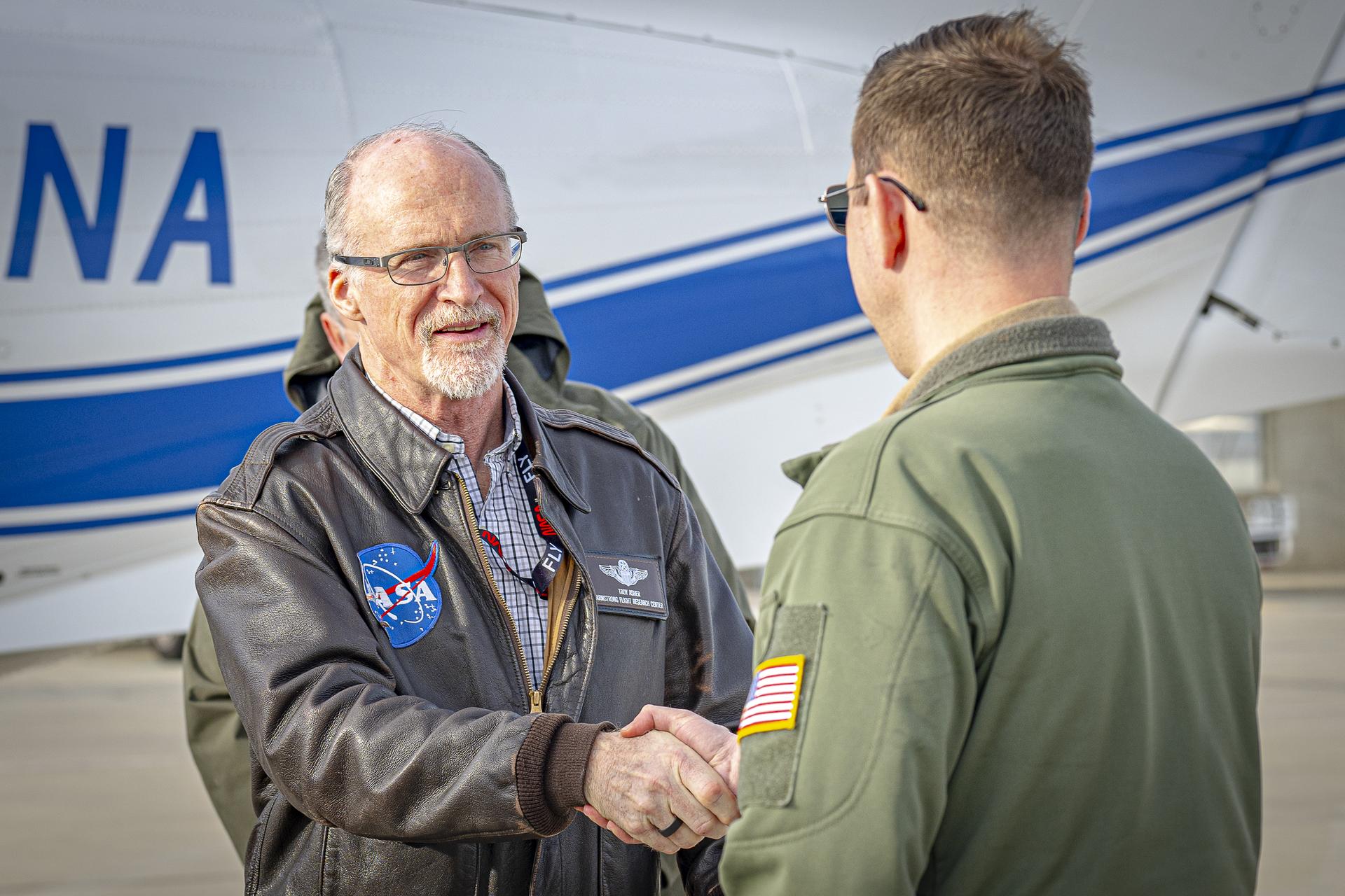 Two men stand facing each other, one in a green flight jacket, and one in a brown flight jacket. They shake hands while standing in front of the back of a white aircraft with a blue stripe.