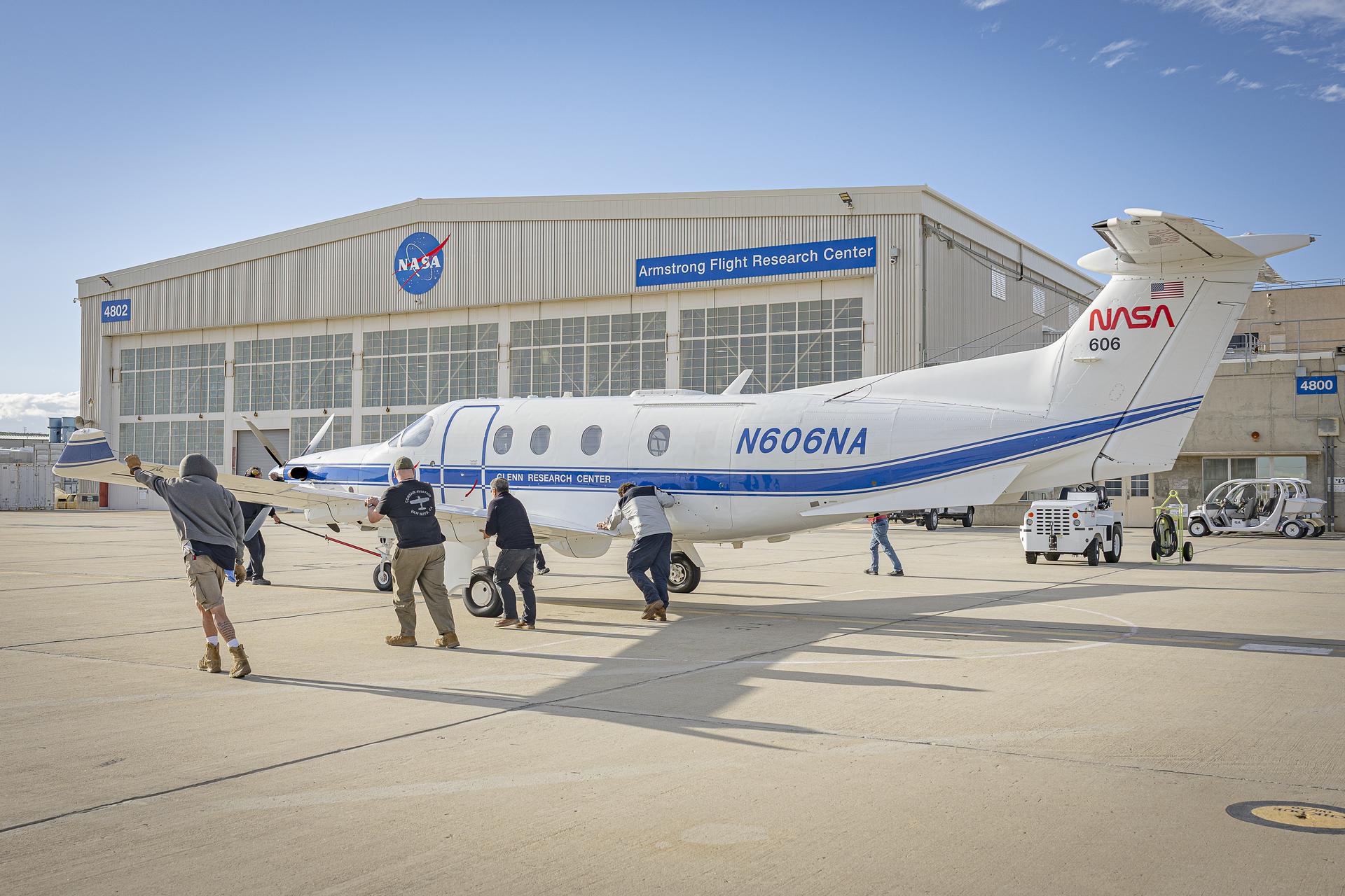 A white aircraft with a front silver propellor is being towed with a red bar and pushed by a crew of workers on a concrete surface. The aircraft is painted with a blue stripe and a round NASA logo on the side and has a red NASA horizontal logo on the tail. A man in a blue jacket and tan pants climbs out of the aircraft carrying two black bags. A silver hangar sits in the background with large white doors. A round NASA logo is located on the hangar as well as a blue sign with white letters.