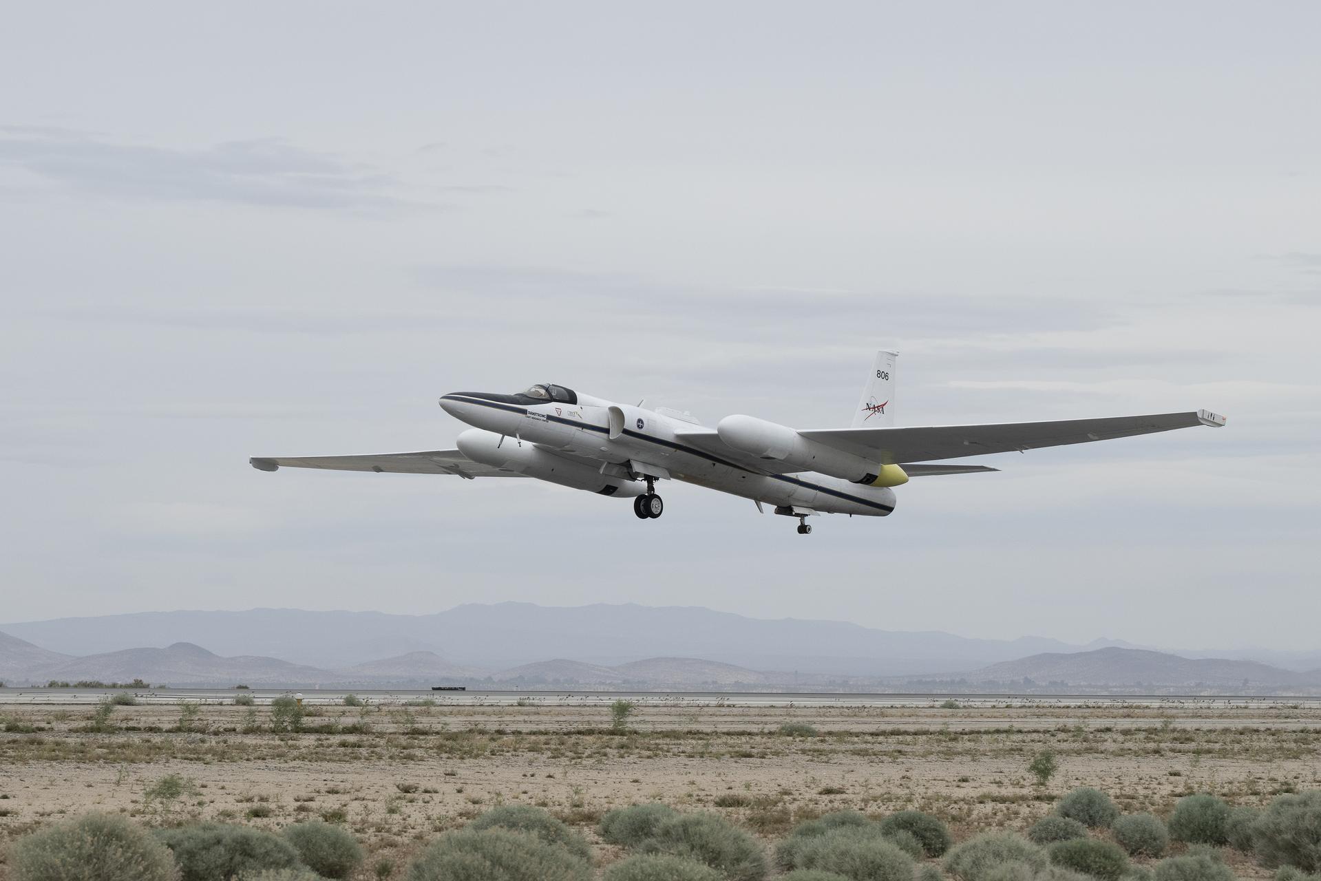 A white aircraft with long, thin wings takes off from a runaway. The ground is below covered in sand and light green, small bushes. There are mountains in the background slightly covered by smoke or fog. The aircraft has two large pods connected to the wings. There are two sets of wheels underneath the aircraft and a small cockpit.