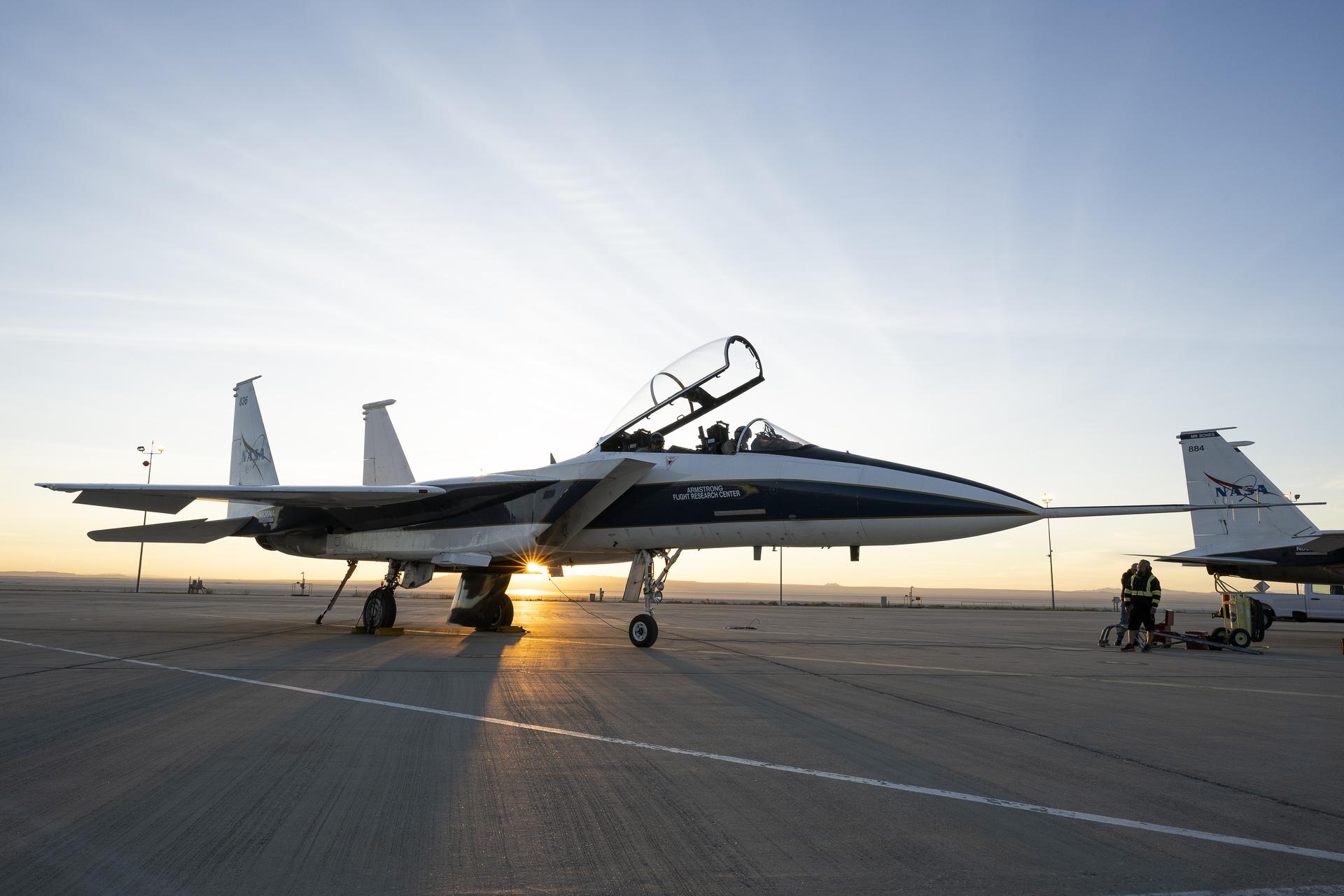 An F-15 research aircraft sits on the ramp at NASA’s Armstrong Flight Research Center in Edwards, California, on Tuesday, March 17, 2026. NASA pilots Jim Less, front seat, and Carrie Worth prepare for the flight. The agency’s Crossflow Attenuated Natural Laminar Flow (CATNLF) test article is attached to the bottom of the F-15. The project aims to lower fuel costs for future commercial aircraft by testing a scale-model wing designed to improve laminar flow.