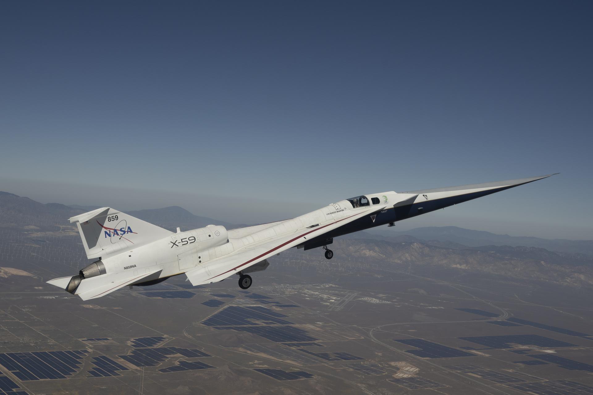 NASA’s X-59 quiet supersonic research aircraft flies above the Mojave Desert on a clear day. The aircraft is white with light gray, red, and blue accent colors. A NASA logo is visible on its tail, along with the number 859 above it.