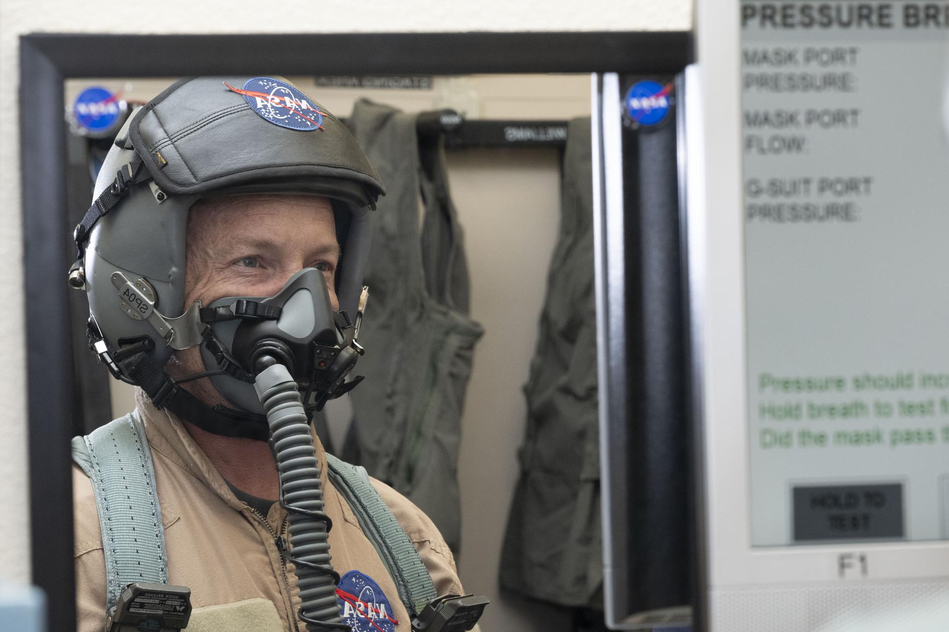 A man wears a helmet with the NASA logo and face mask with a tube coming down that connects to a tank. He is wearing a tan flight suit with the NASA logo and green harness straps.