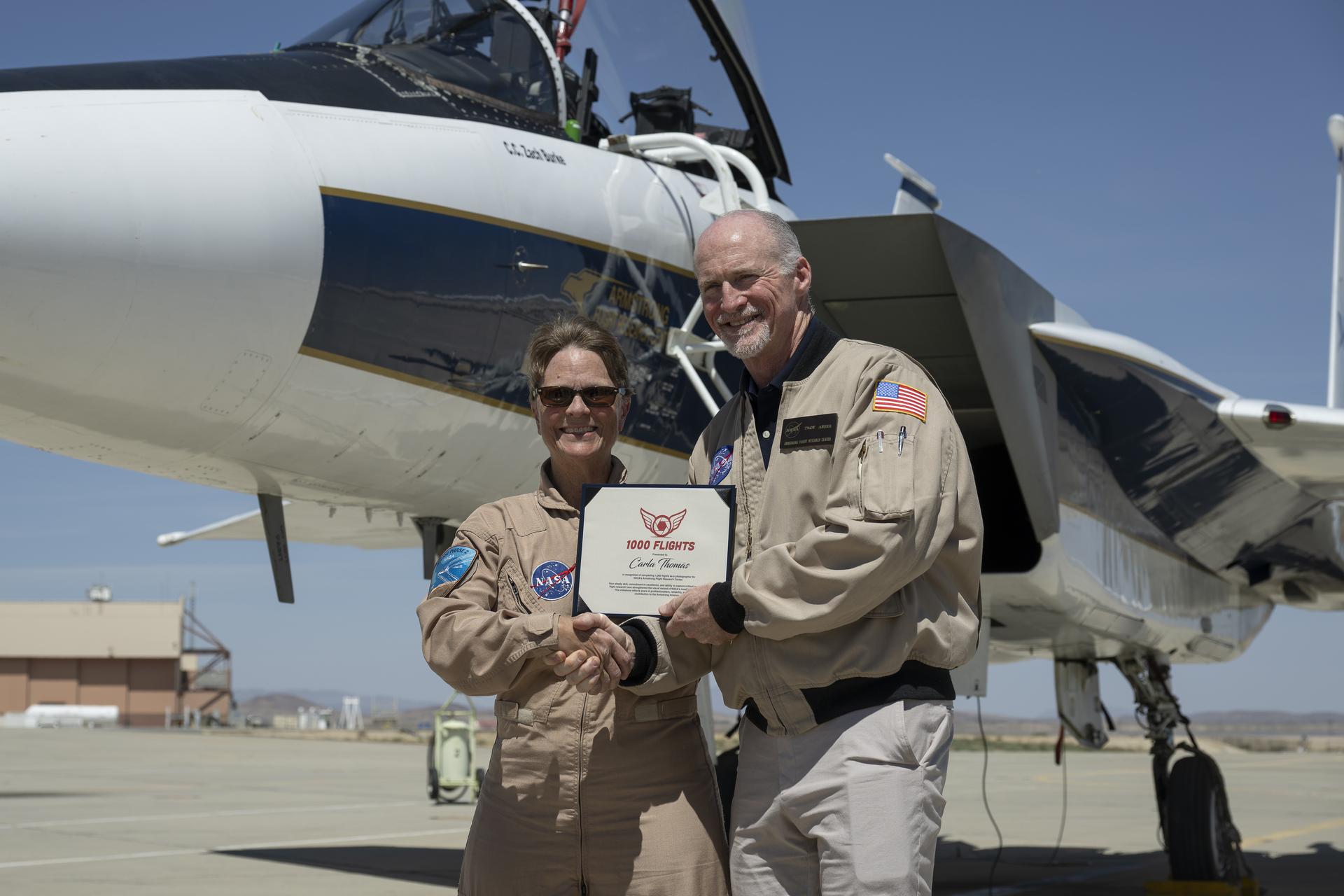 Troy Asher, acting center director, presents NASA aerial photographer Carla Thomas with an award commemorating her 1,000th flight at NASA’s Armstrong Flight Research Center in Edwards, California, on Monday, April 20, 2026. For 38 years, Thomas has captured aerial photographs of research and science flights. The milestone flight was completed in the backseat of a NASA F-15 aircraft piloted by NASA Armstrong chief pilot Wayne Ringelberg.