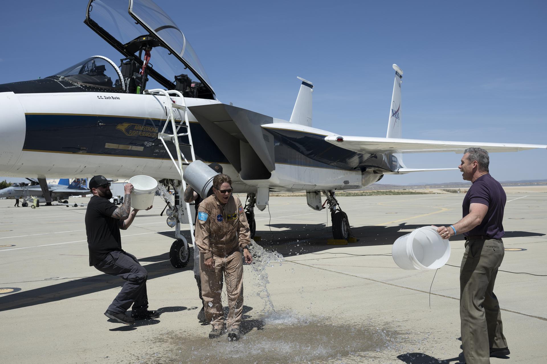 F-15 aircraft crew chief Zack Burke, left, and X-59 mechanic Jamie Trescott, center back, splash buckets of ice water on NASA aerial photographer Carla Thomas in celebration of her 1,000th flight at NASA’s Armstrong Flight Research Center in Edwards, California, on Monday, April 20, 2026. Tim Krall, NASA Armstrong support aircraft fleet manager, watches. Thomas completed her flight in the backseat of a NASA F-15 aircraft flown by NASA Armstrong chief pilot Wayne Ringelberg. For 38 years, Thomas has captured aerial photographs of research and science flights.