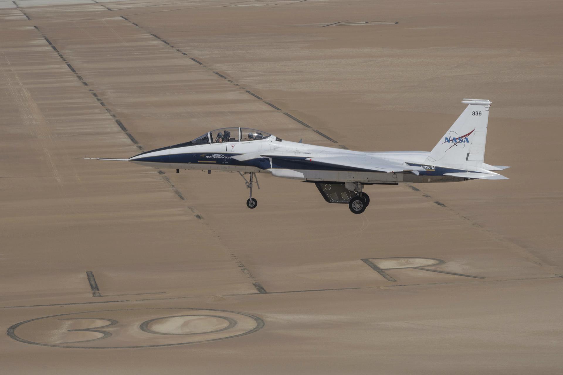 A white and blue painted jet aircraft flies above a tan lakebed with the gear and wheels out. A model wing hangs below the aircraft’s center line. The cockpit is closed and two pilots are visible inside with flight gear on.