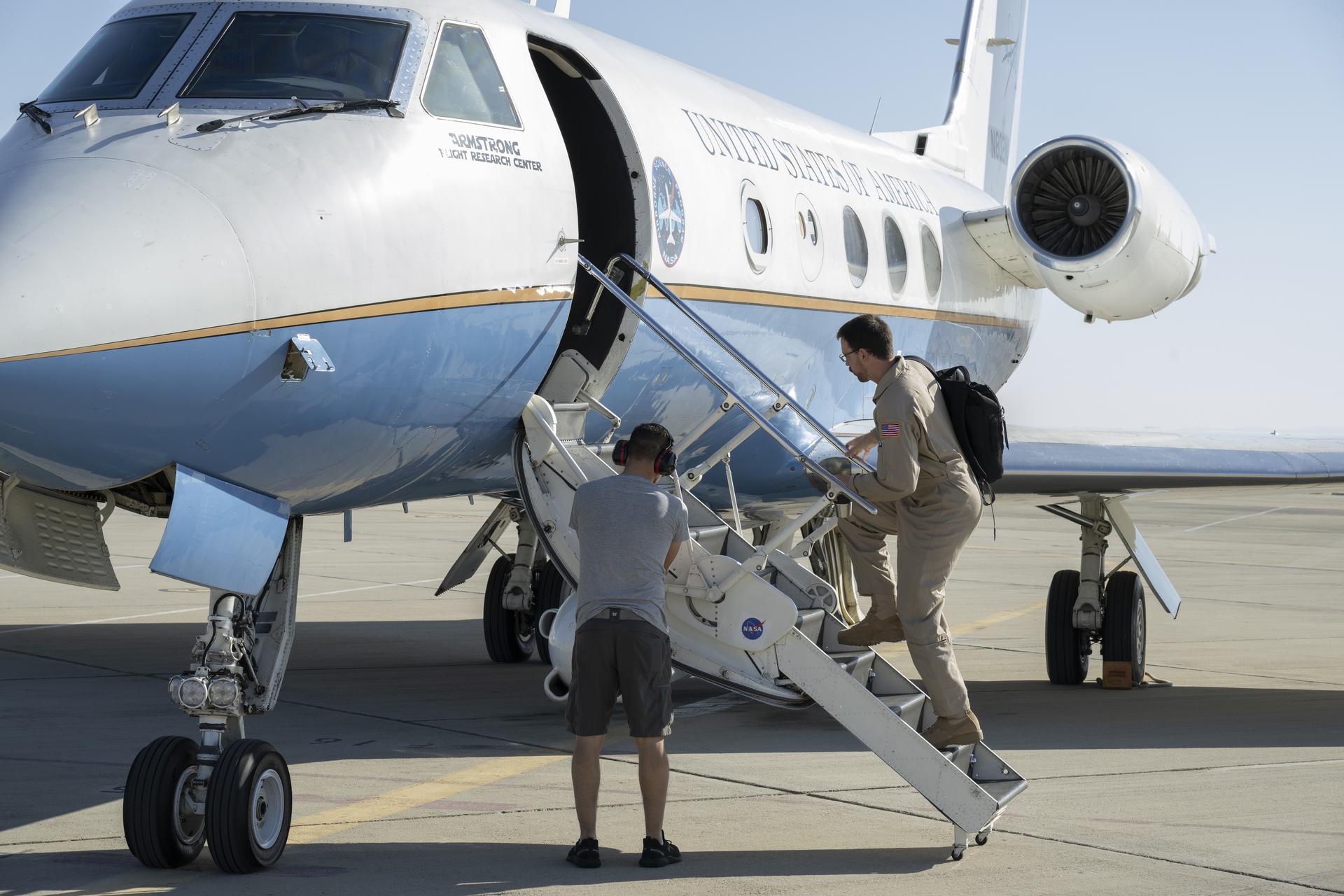 A man stows equipment in a compartment of the aircraft stairs, while another man goes up the stairs to get into the aircraft.