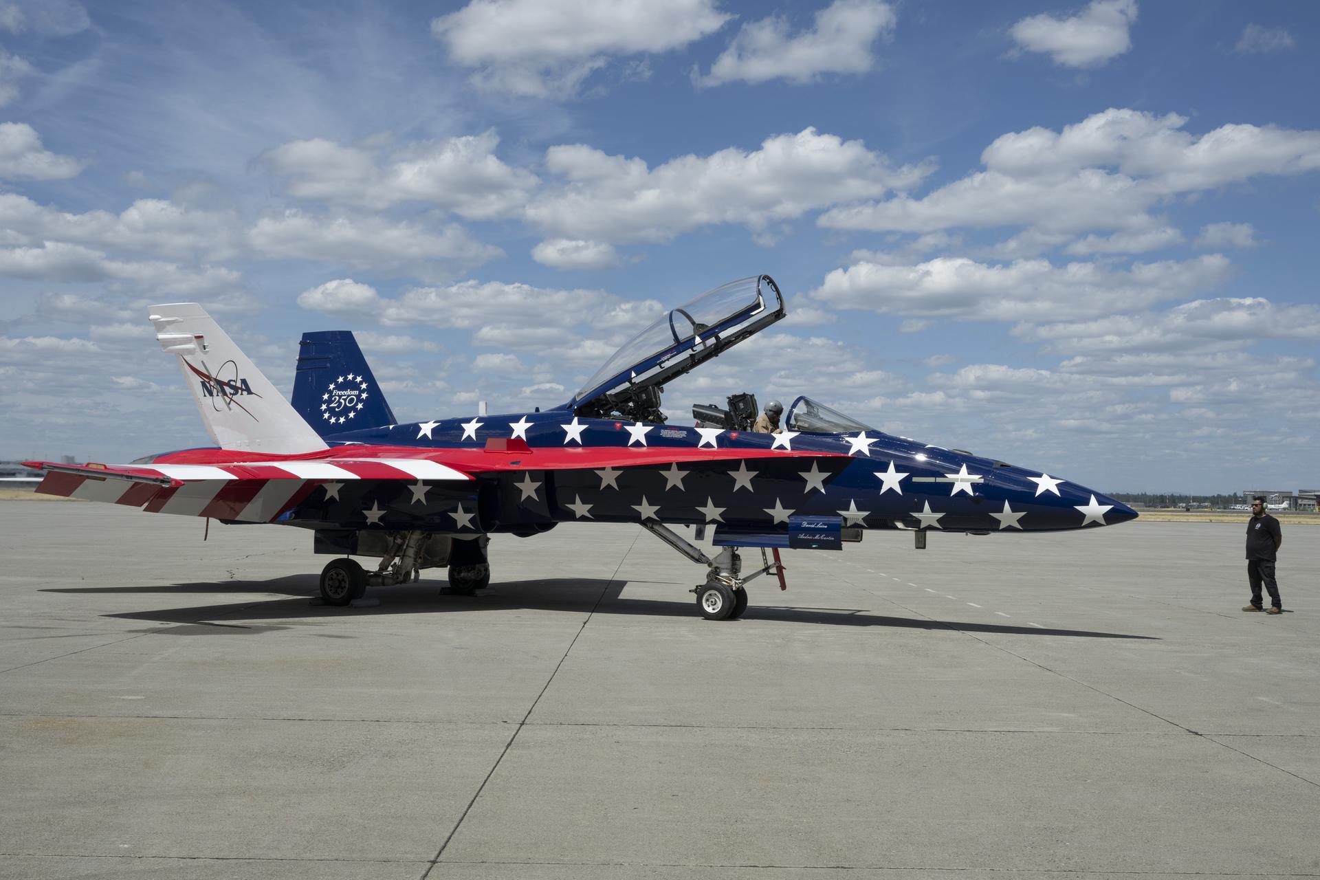 A pilot sits inside a red, white, and blue jet aircraft on the ramp. Another person wearing headphones stands in front of the aircraft. The aircraft’s body is painted blue with white stars, and its wings feature red and white stripes resembling the American flag.