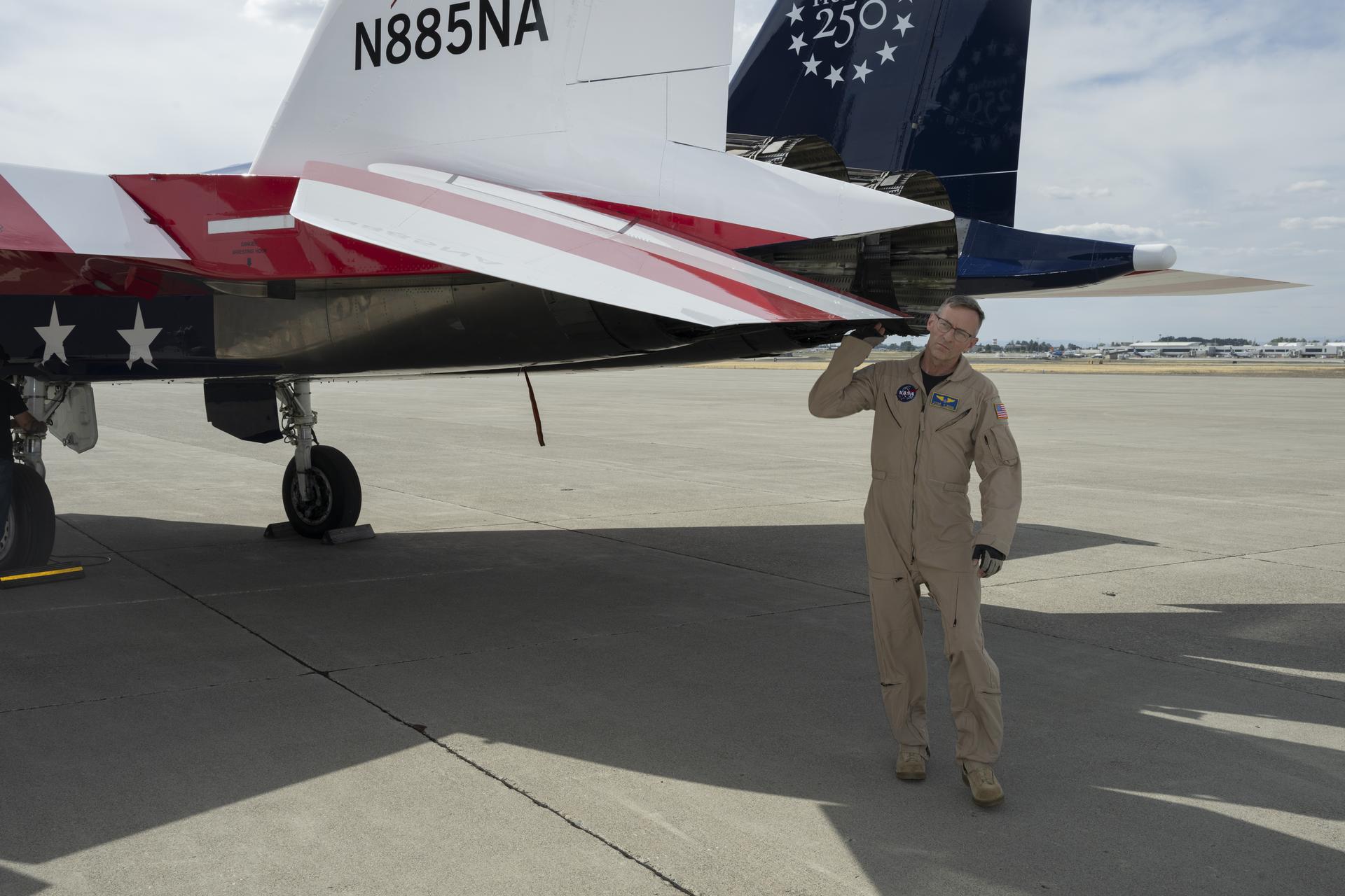 A pilot stands beside a red, white, and blue jet aircraft on the ramp, performing pre‑flight checks. The aircraft’s body is painted blue with white stars, and its wings have red and white stripes.