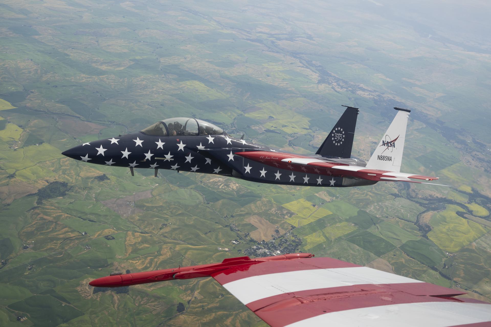 A red, white, and blue NASA F‑15 is shown beside another jet aircraft during a cross‑country flight. The full F‑15 is visible, while only a portion of the F/A‑18’s red‑and‑white wing appears in the frame.