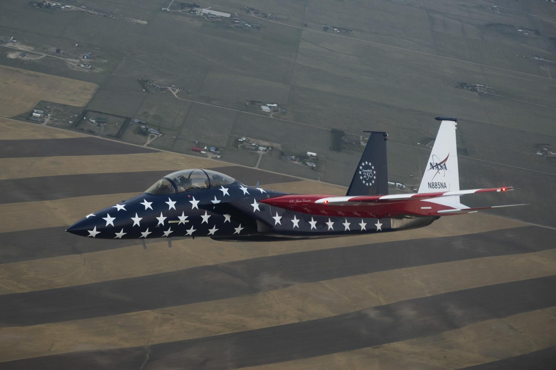 A red, white, and blue jet aircraft is shown during a cross‑country flight. The body of the aircraft is painted blue with white stars, and the wings are red and white stripes resembling the American flag.