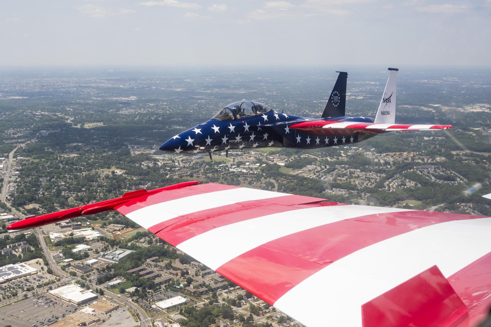 A NASA F-15 aircraft flies above Washington on Saturday, July 4, 2026, as part of a flyover to celebrate America’s 250th birthday. The wing of a NASA F-18 is seen in the foreground, as a NASA photographer captured this image from inside the cockpit. This aircraft is from NASA’s Armstrong Flight Research Center in Edwards, California, and it joined other NASA aircraft for the flyover.