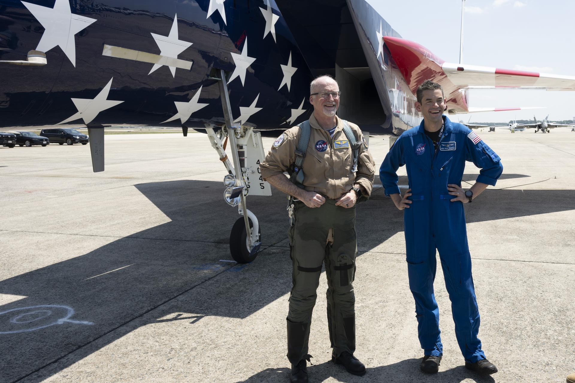 Two men stand in front of a red, white, and blue painted jet aircraft. The man on the right wears a blue flight suit and the man on the left wears a tan flight suit.