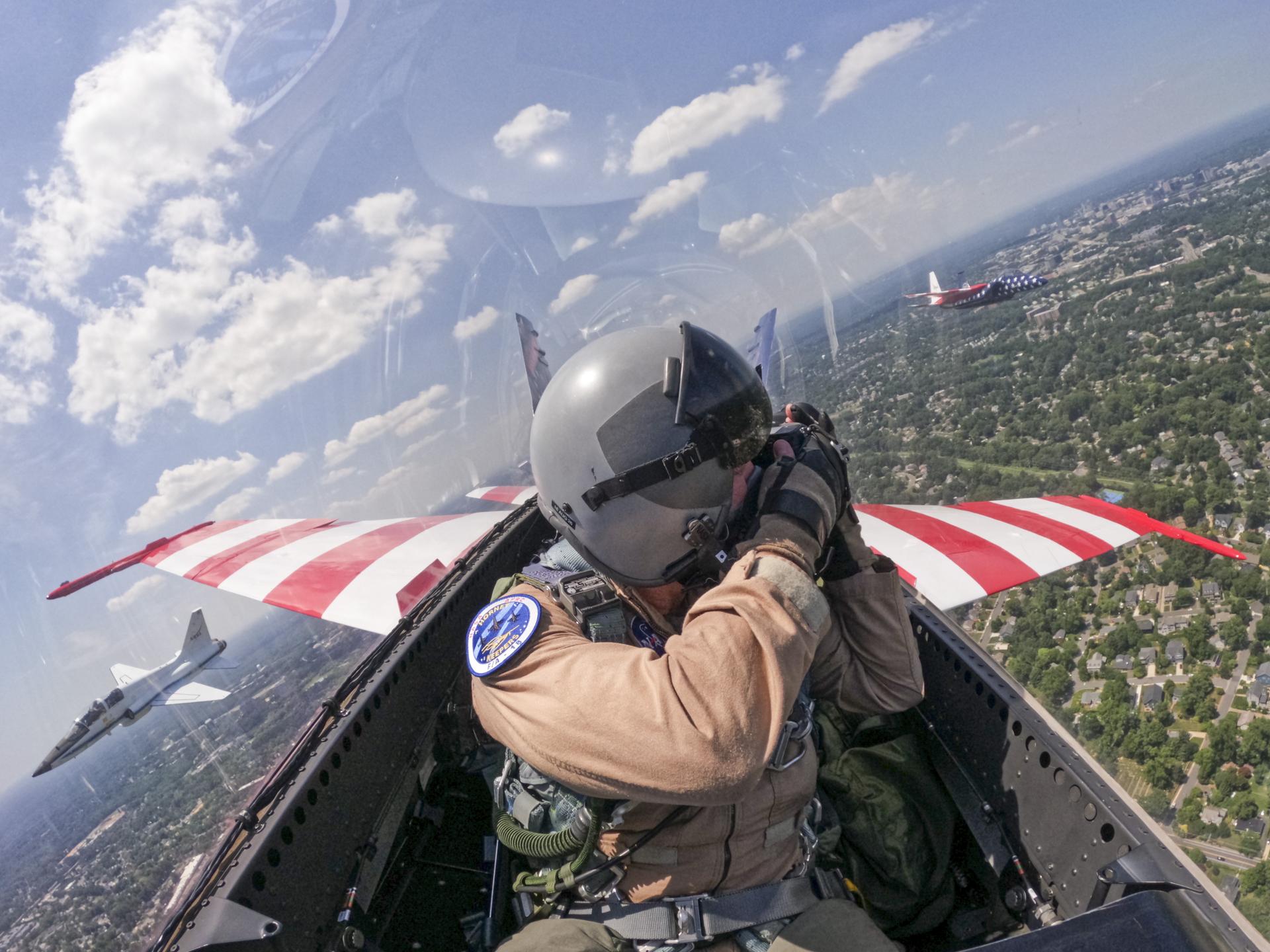 A man in a tan flight suit, flight gear, and helmet looks outside of the jet aircraft cockpit windows with a camera. Two other jet aircraft are seen outside of the cockpit window.