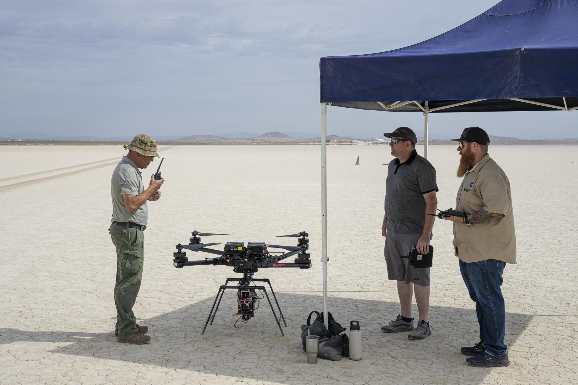 One man communicates with a hand-held radio, as two other men wait to the side for the go-ahead command to fly an experiment on a drone.