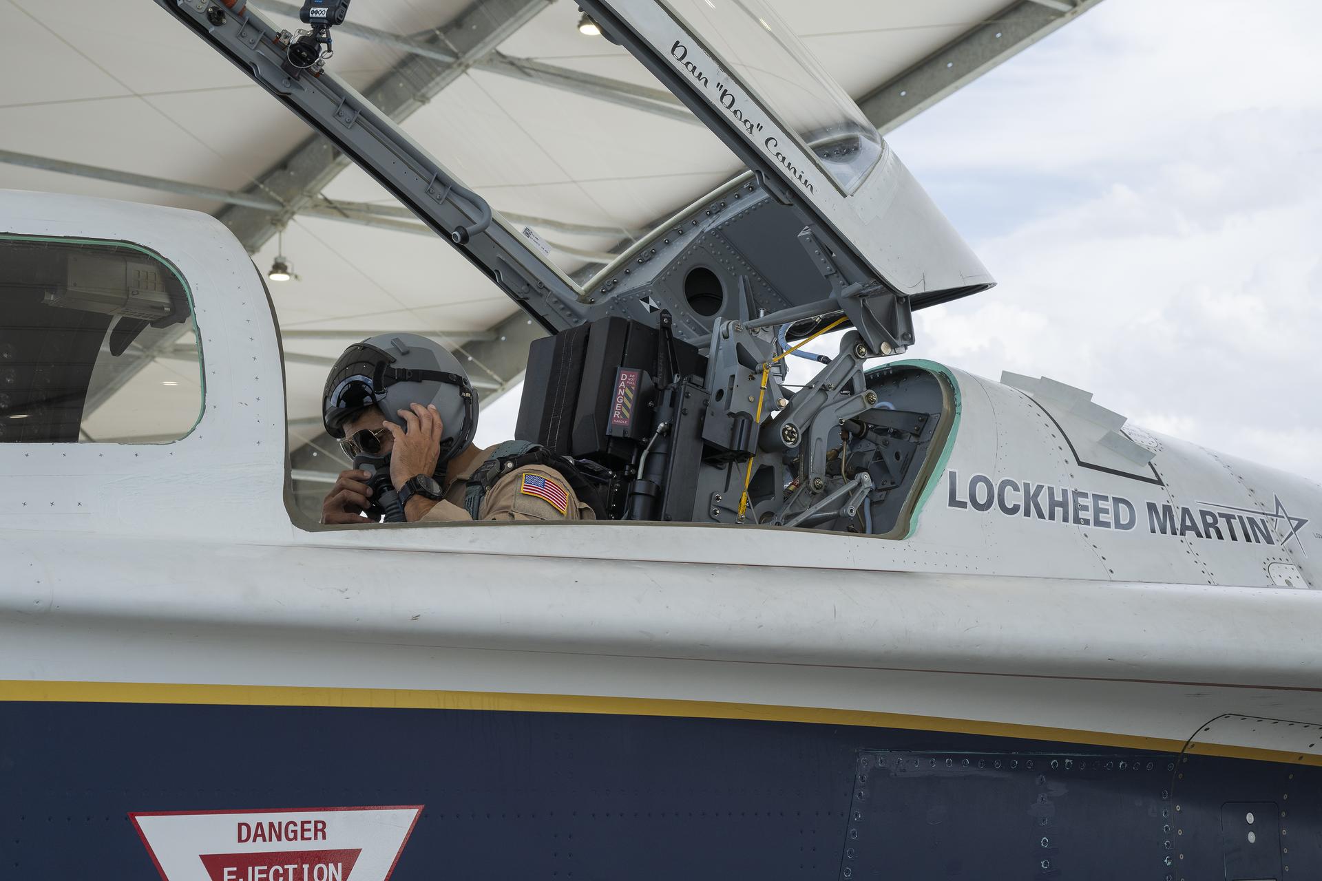 A man seated inside an aircraft cockpit adjusts a helmet, with the open canopy above him.