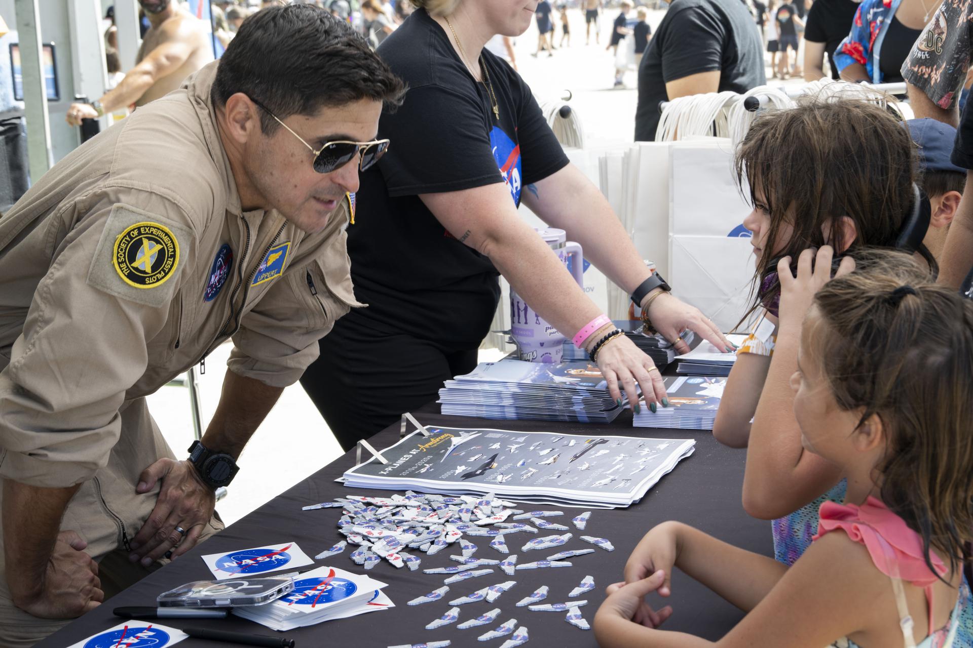 NASA test pilot Mike Lippert talks with young visitors at the NASA exhibit during the Pacific Airshow at Huntington Beach, California, Friday, Oct. 2, 2026. The exhibit features Artemis, the X‑59, SkyFall, a lunar globe, and an Apollo 15 Moon rock.
