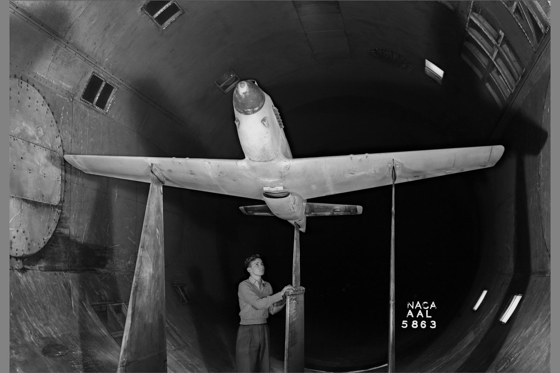 An airplane sits on a supporting strut in a wind tunnel as a technician looks on.