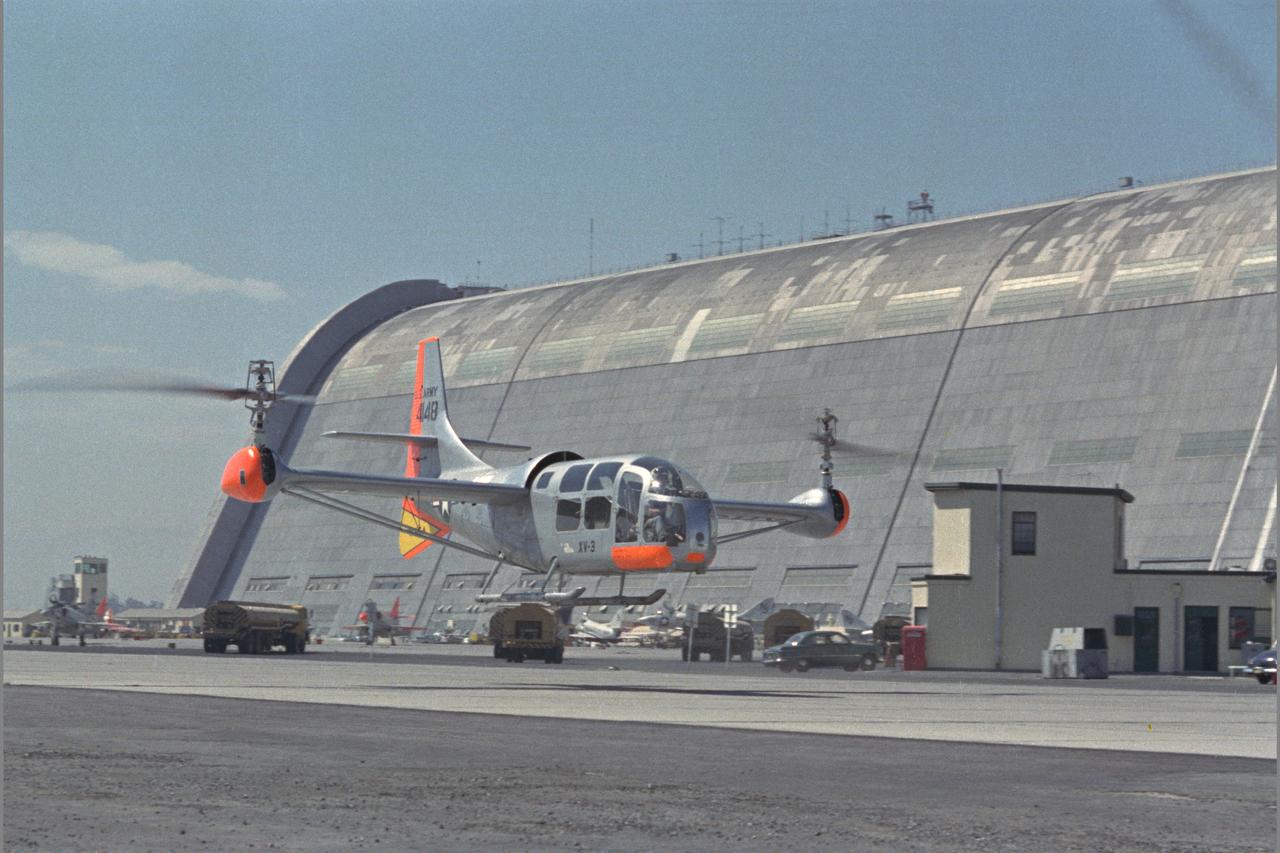 XV-3 HOVERING ON RAMP. Flight Test of Bell XV-3 Convertiplane. Bell VTOL tilt-rotor aircraft hovering along side Hangar One at Moffett Field. The XV-3 design combined a helicopter rotor and a wing. A 450 horsepower Pratt & Whitney piston engine drove the two rotors. The XV-3, first flown in 1955 , was the first tilt-rotor to achieve 100% tilting of rotors. The vehicle was underpowered, however, and could not hover out of ground effect. Note the large ventral fin, which was added to imrpove directional stability in cruse (Oct 1962)