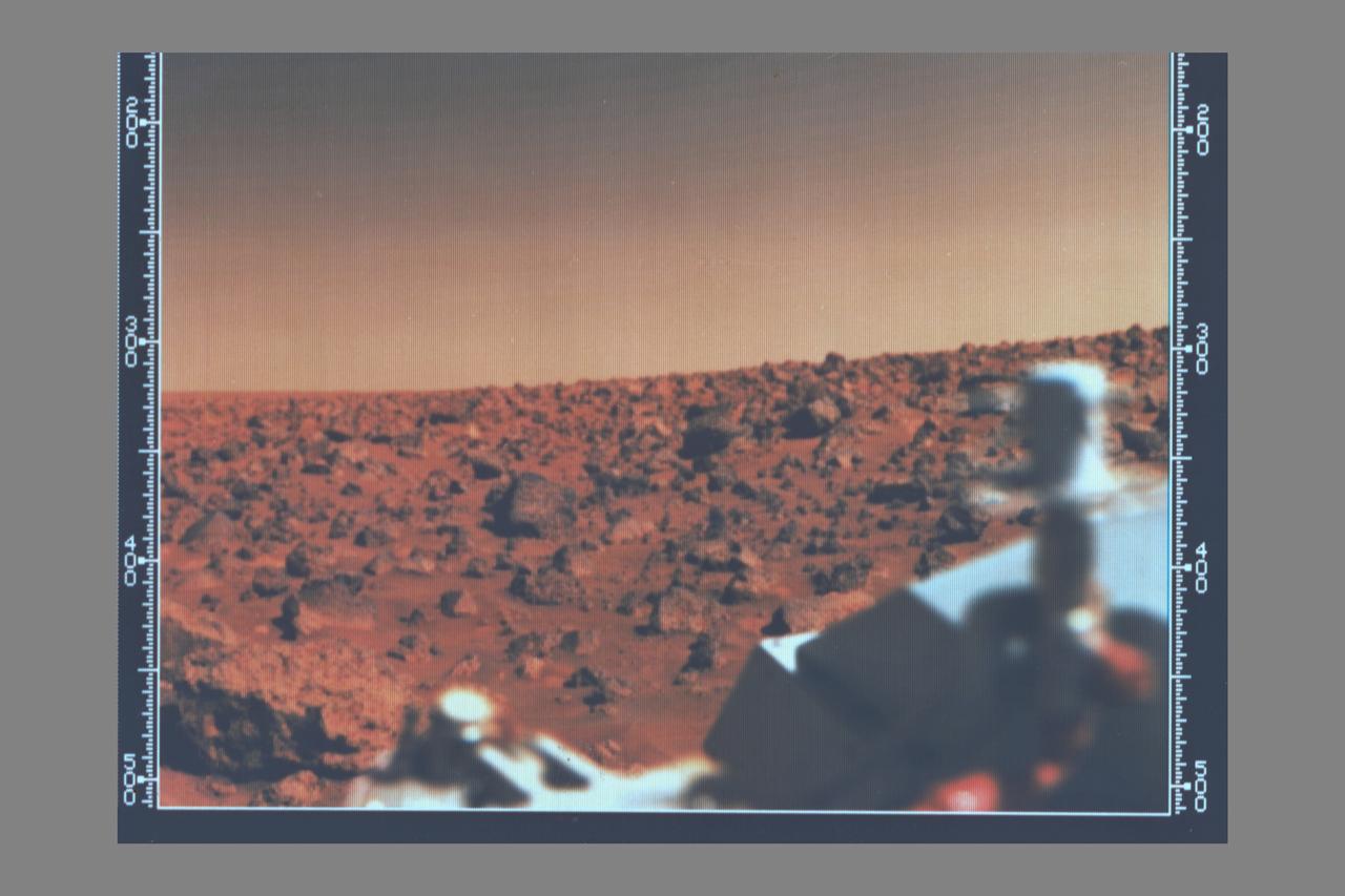 Viking 2 A utopian bright summer afternoon on Mars -- Looking south from Viking 2 on September 7, 1976 the orange-red surface of the nearly level plain upon which the spacecraft sits is seen strewn with rocks as large as three feet across.  Many of these rocks are porous and sponge-like, similar to some of Earth's volcanic rocks.  Other rocks are coarse-grained such as the large rock at lower left.  Between the rocks, the surface is blanketed with fine-grained materials that, in places, is piled into small drifts and banked against some of the larger blocks.  The cylindrical mast with the orange cable is the low-gain antenna used to receive cammands form Earth.  (JPL ref: P-17690 color)